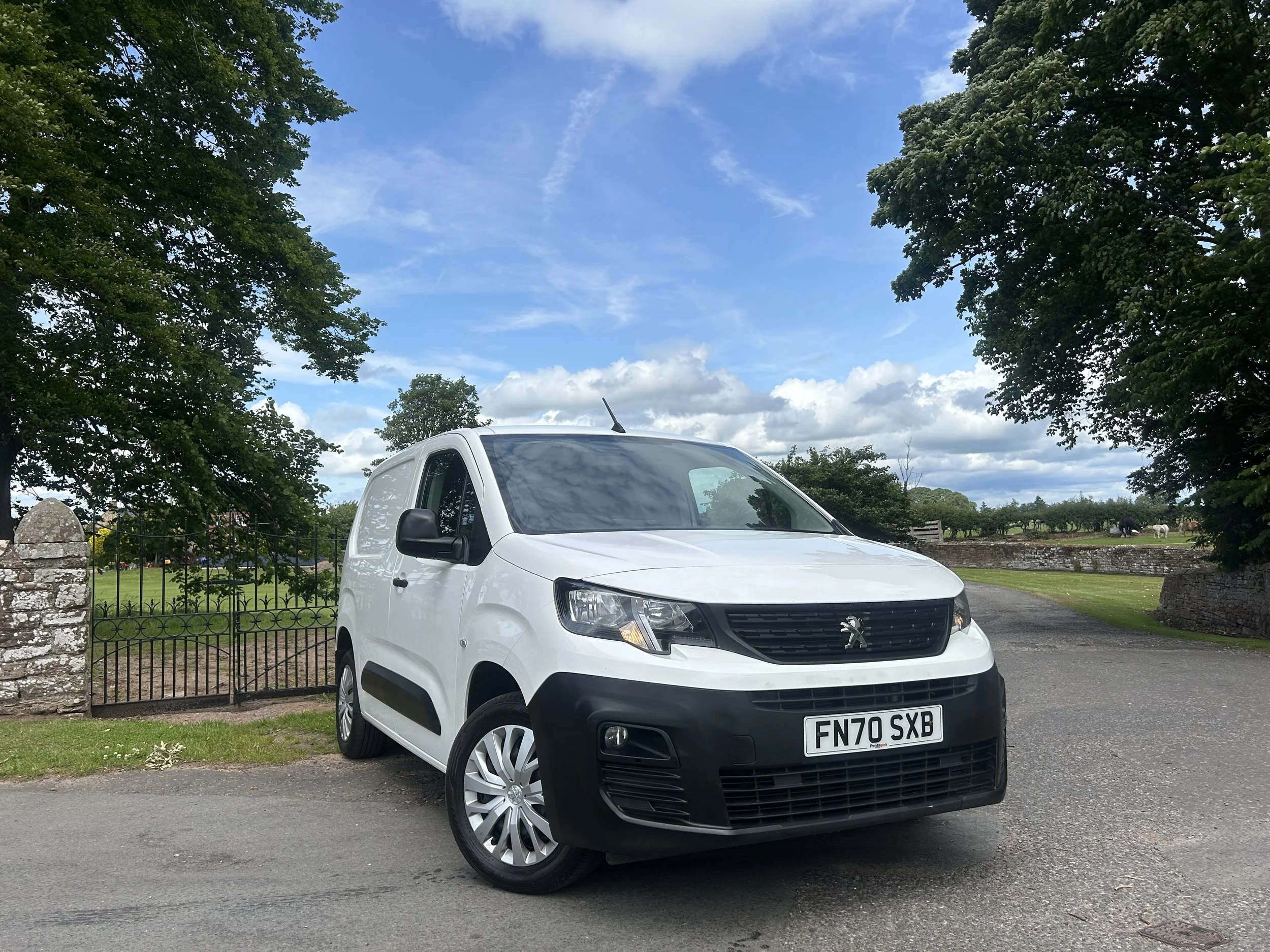 White Peugeot small cargo van parked on a paved area near a stone wall, with trees and a field in the background under a partly cloudy sky.