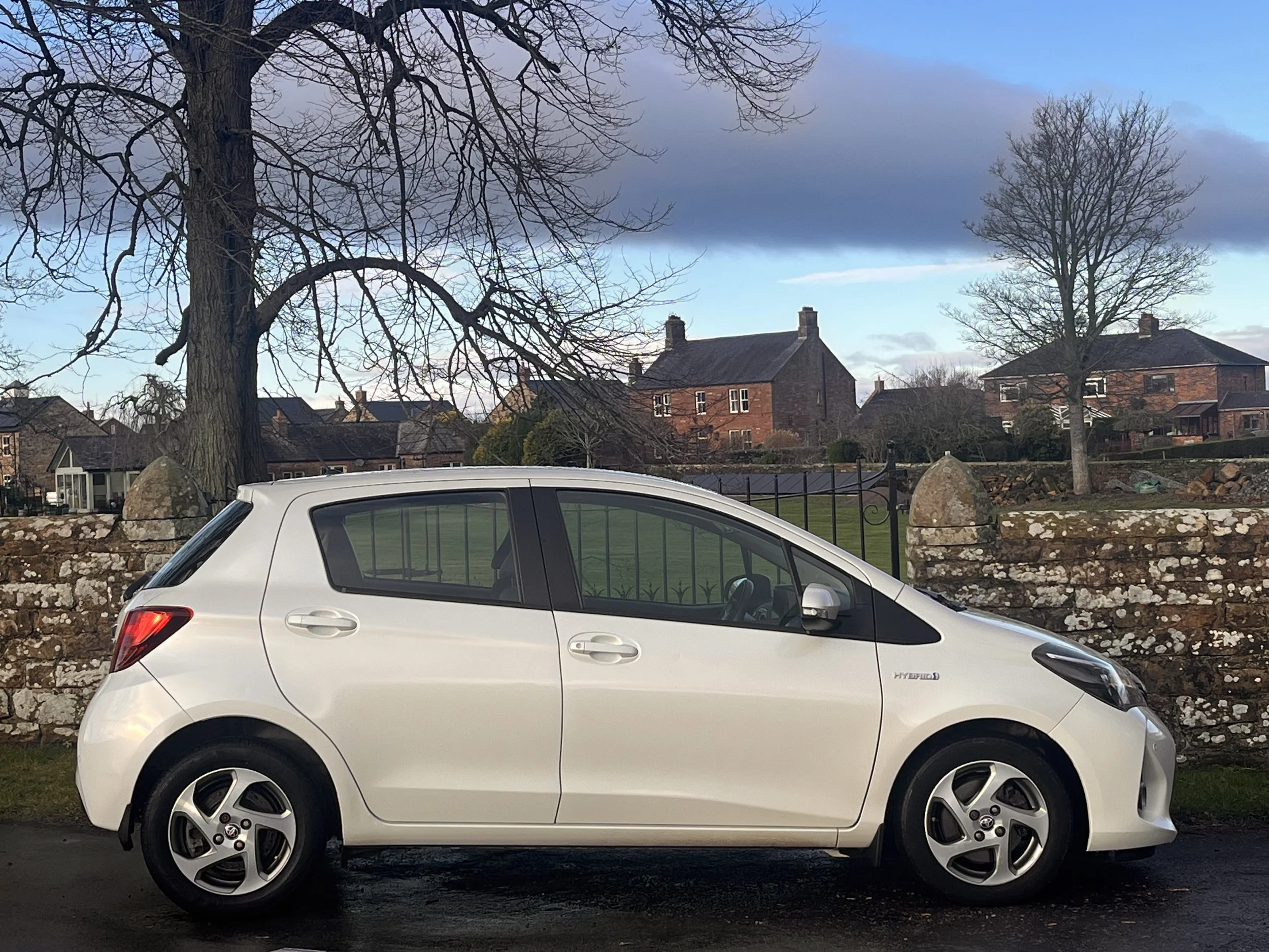 A white hybrid compact car parked on a street with a stone wall, leafless trees, and brick houses in the background under a partly cloudy sky.