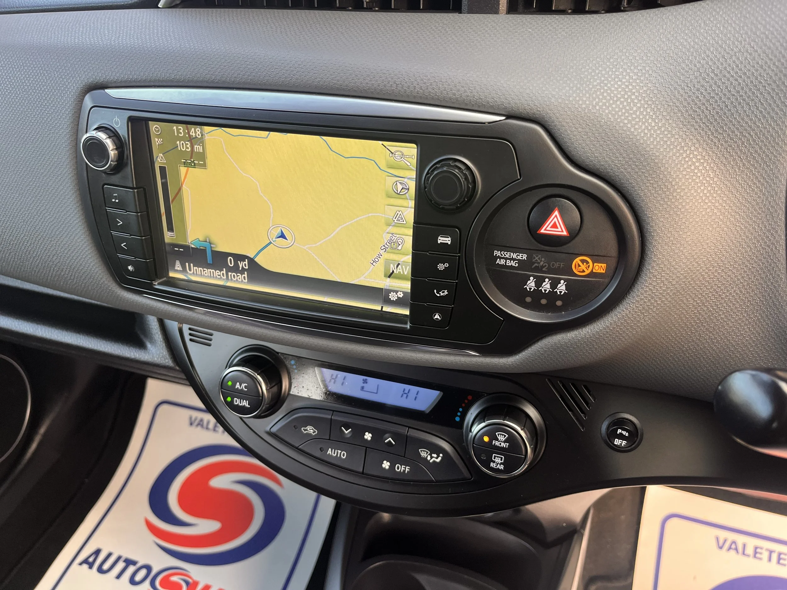 Car dashboard displaying navigation map with the route on an unnamed road, climate control settings, hazard lights button, and passenger airbag indicator.