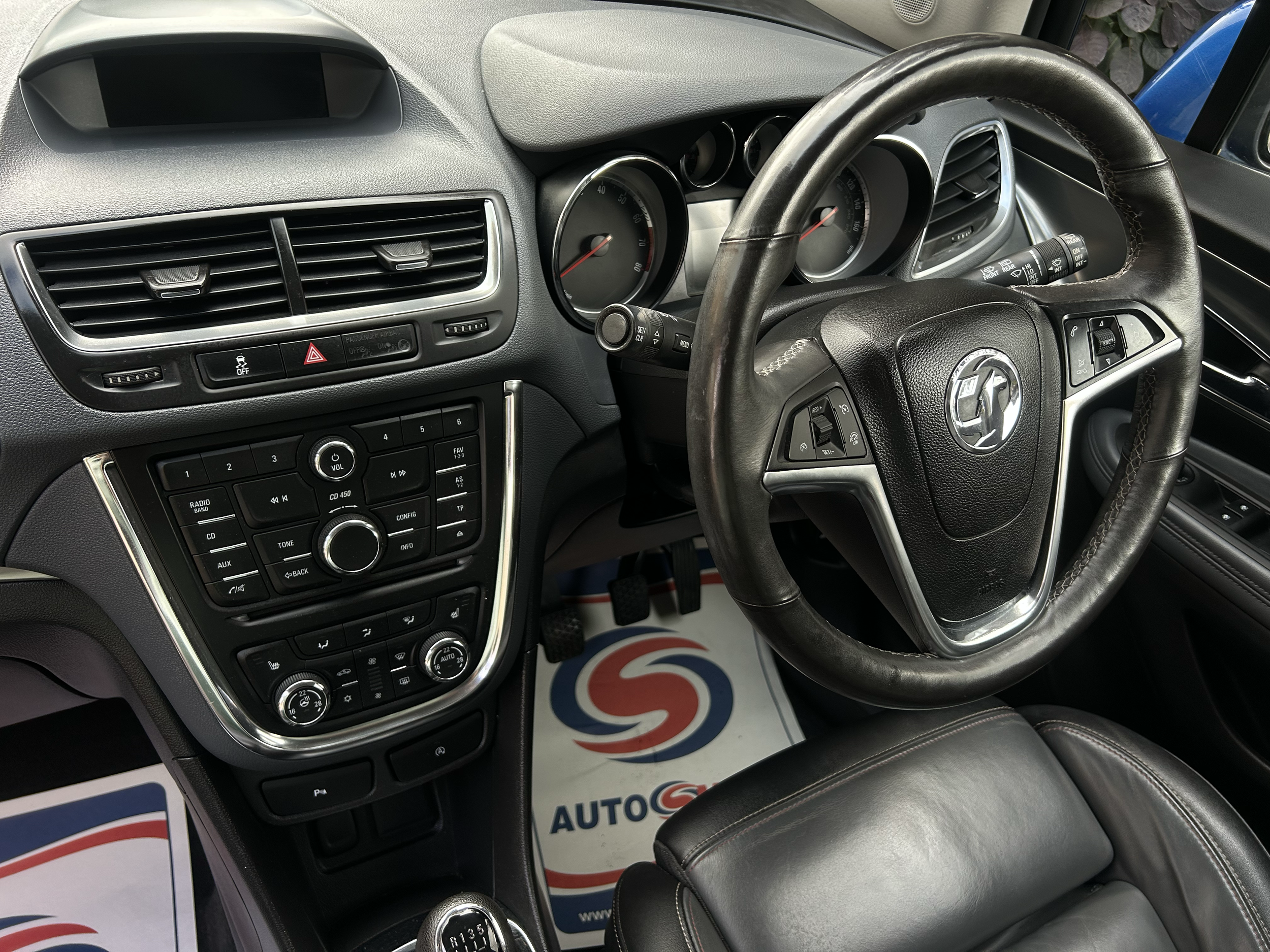 The interior of a vehicle showing a black leather steering wheel, a dashboard with circular gauges, an air vent, and a control panel with buttons and dials, with part of a printed sign or paper in the background.