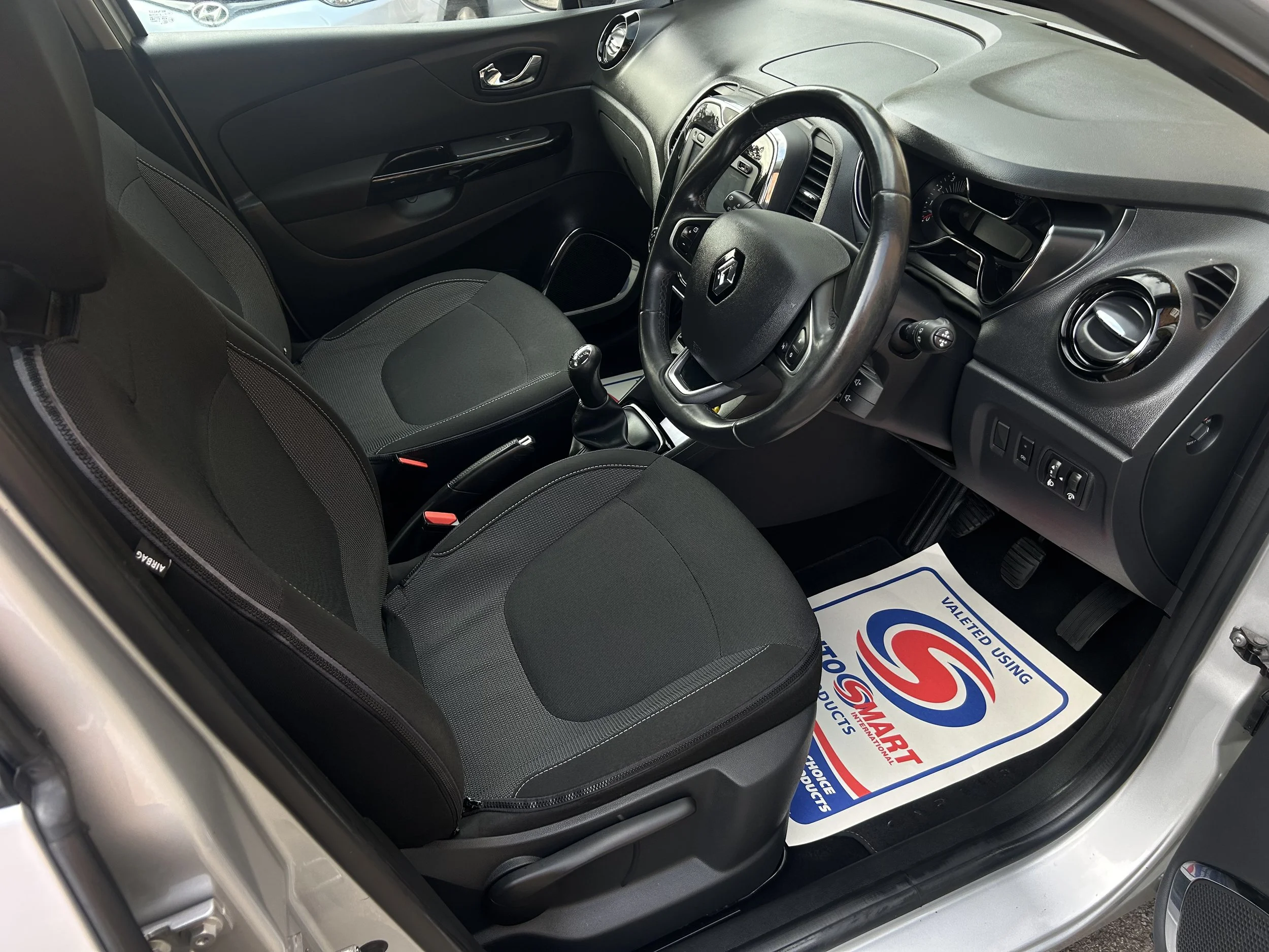 Interior of a car showing the front seats, steering wheel, dashboard, and floor mat with a logo.