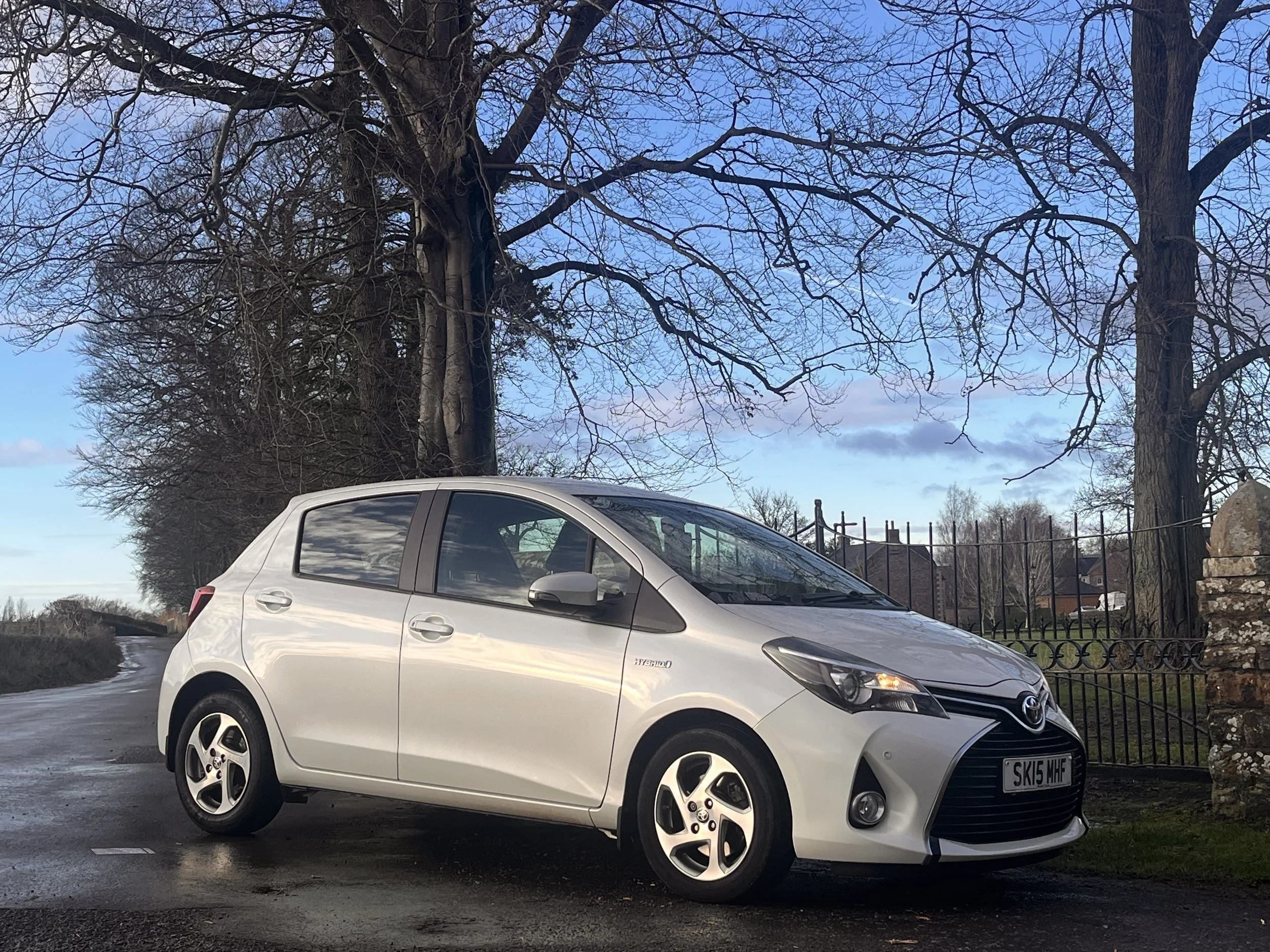 A white Toyota Yaris hybrid parked on a wet street next to a stone wall and black metal fence, with leafless trees and a blue sky in the background.