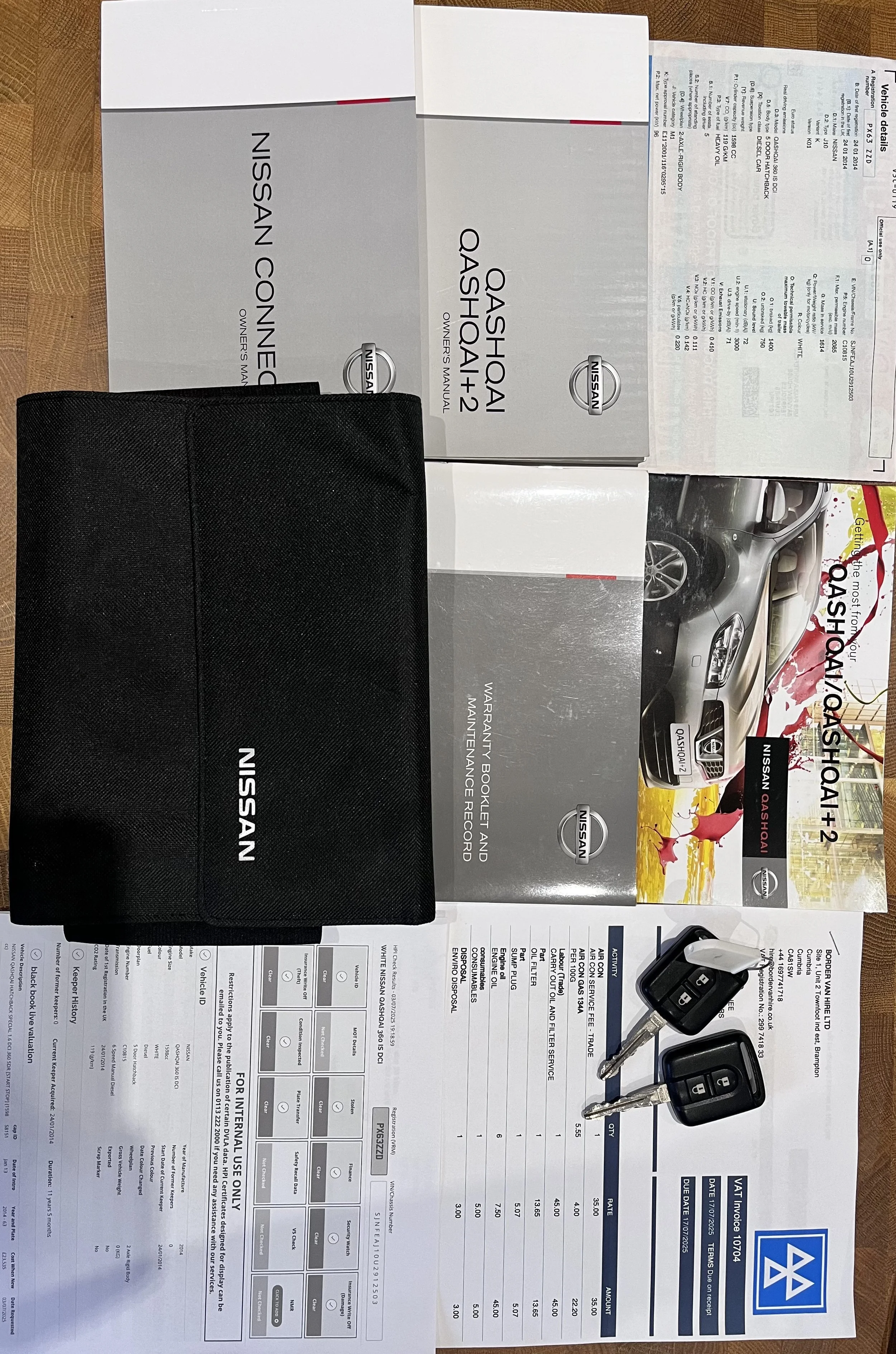 Car keys on a brochure and owner's manuals for a Nissan Qashqai+2, placed on a wooden surface, along with a Nissan-branded black pouch.