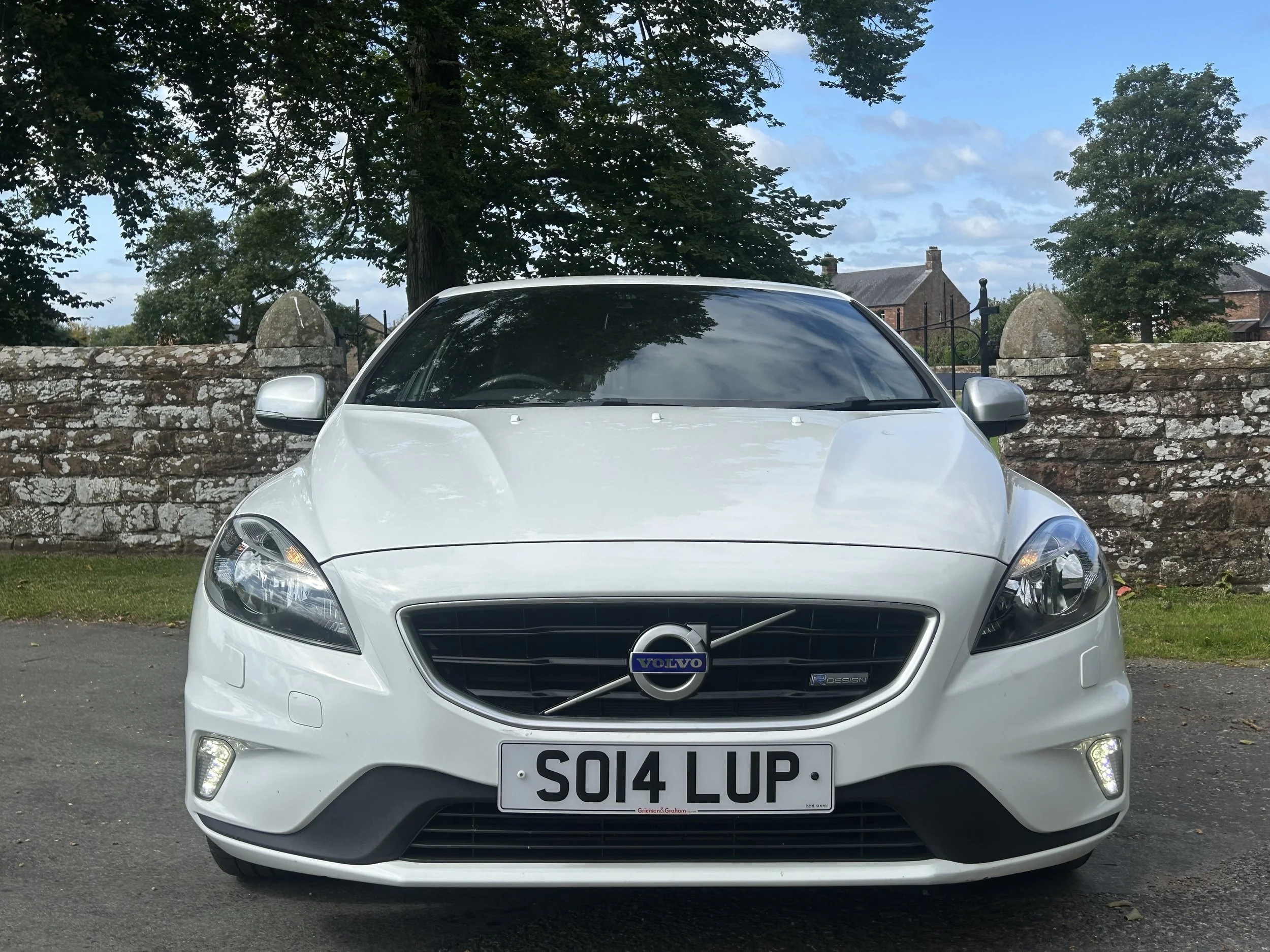 Front view of a white Volvo car parked in front of a stone wall with trees and buildings in the background.