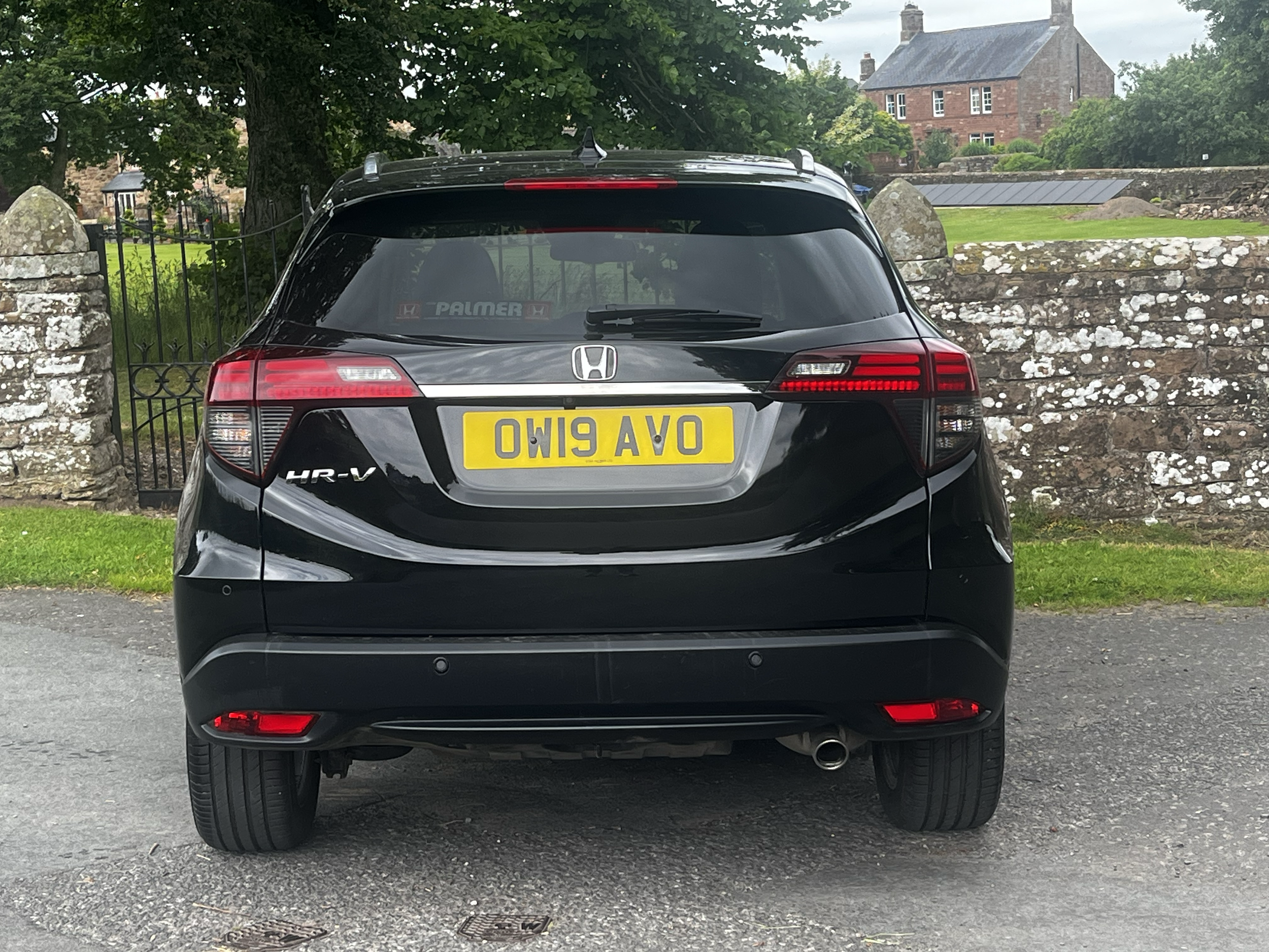 Black Honda HR-V parked on a driveway behind a stone and wrought iron fence, with a rural house and trees in the background under an overcast sky.