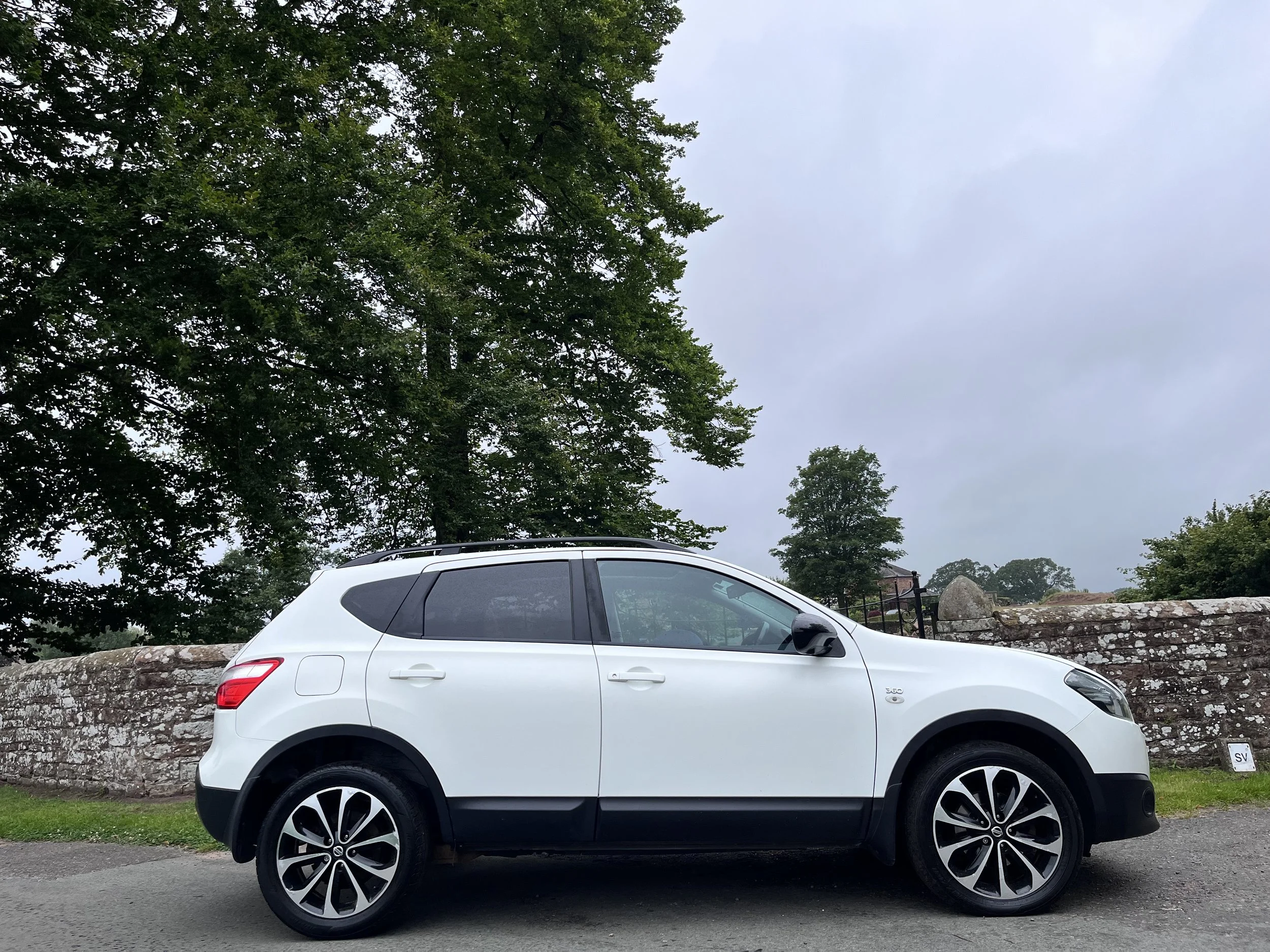 White compact SUV parked on a sidewalk near a stone wall and green trees