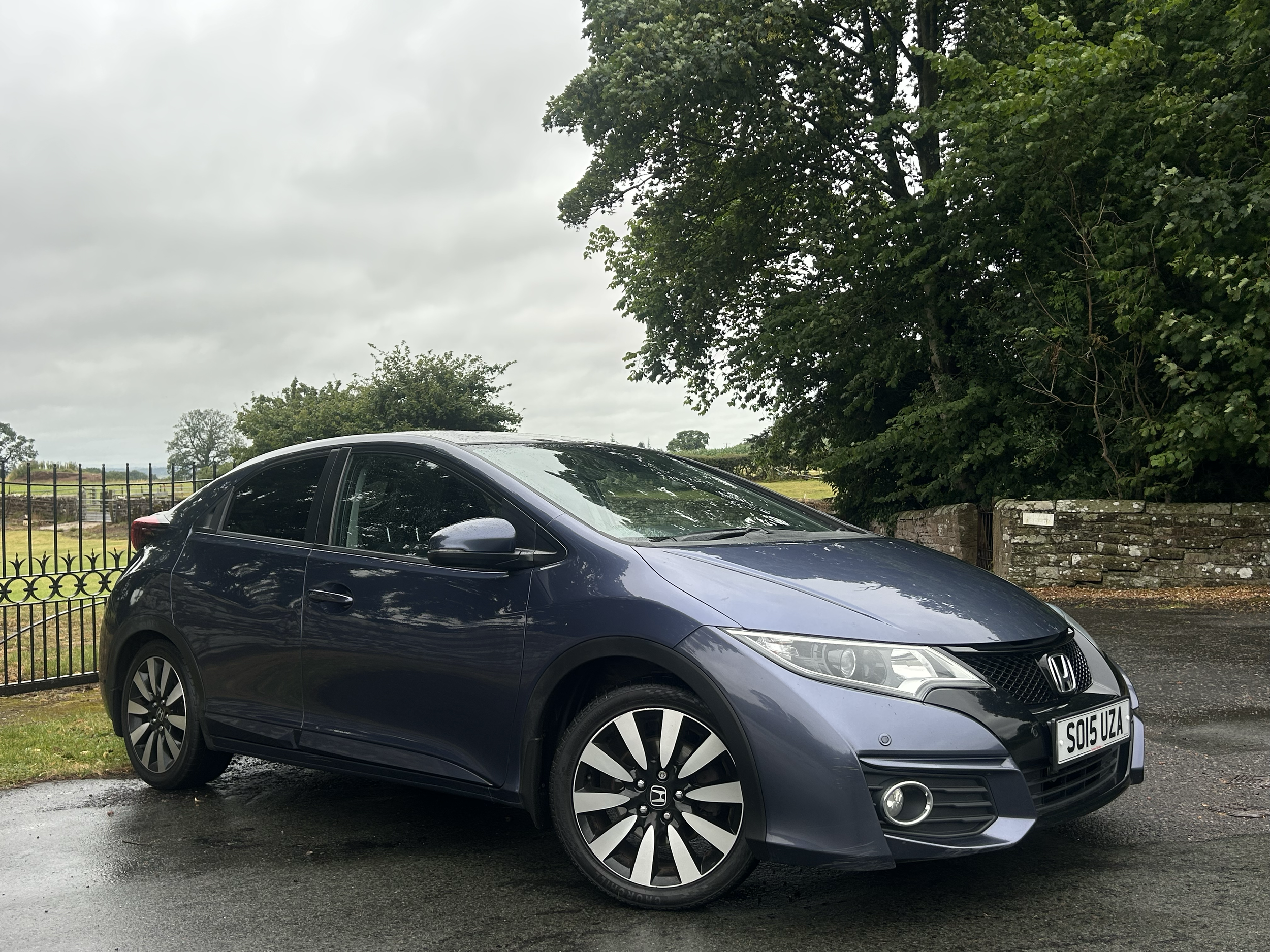 A front view of a dark gray Honda Civic parked on a wet asphalt surface, with large green trees and a cloudy sky in the background.