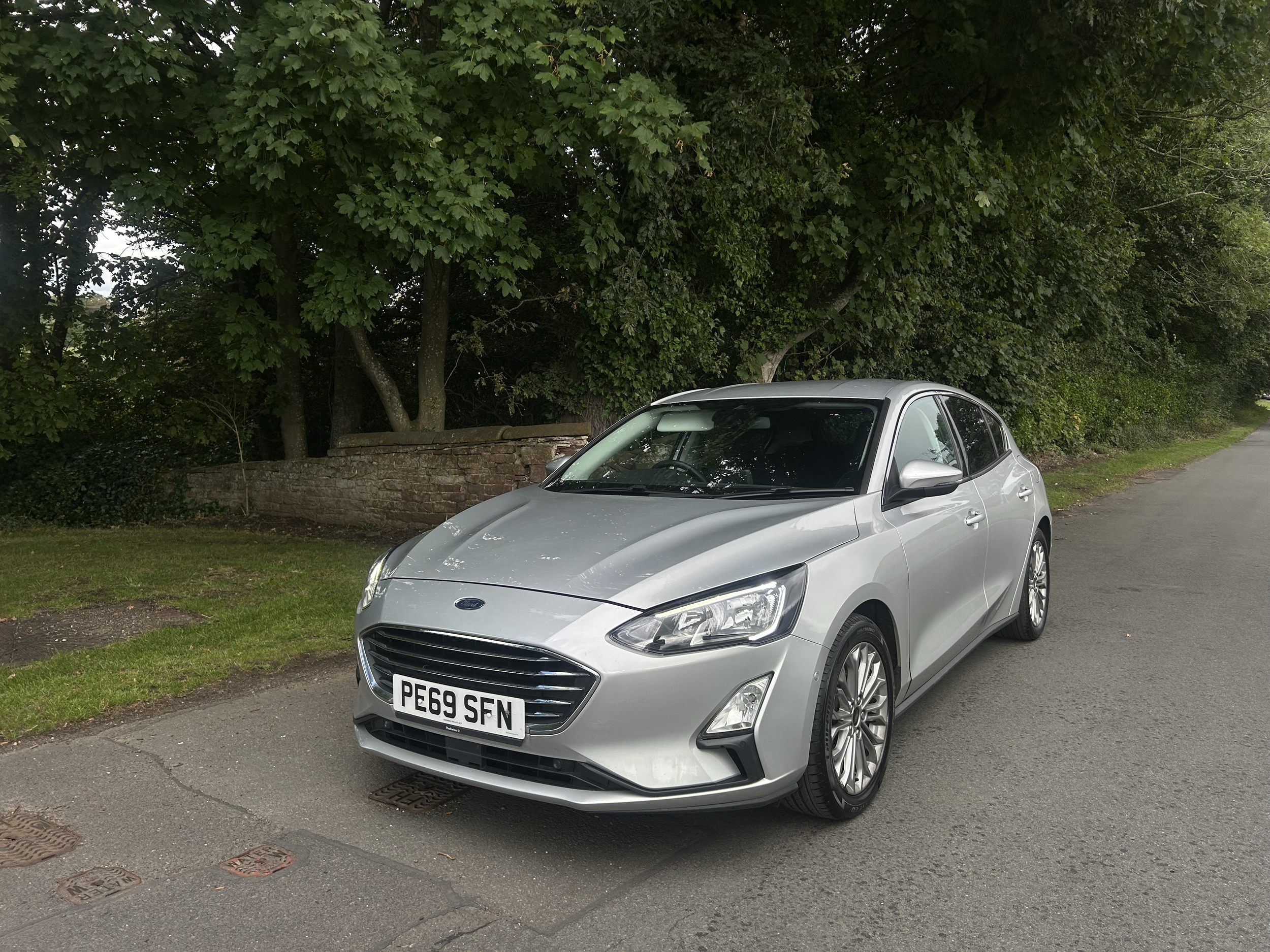 A silver hatchback car parked on the side of a street next to a grassy area and a brick wall, with green trees and bushes in the background.