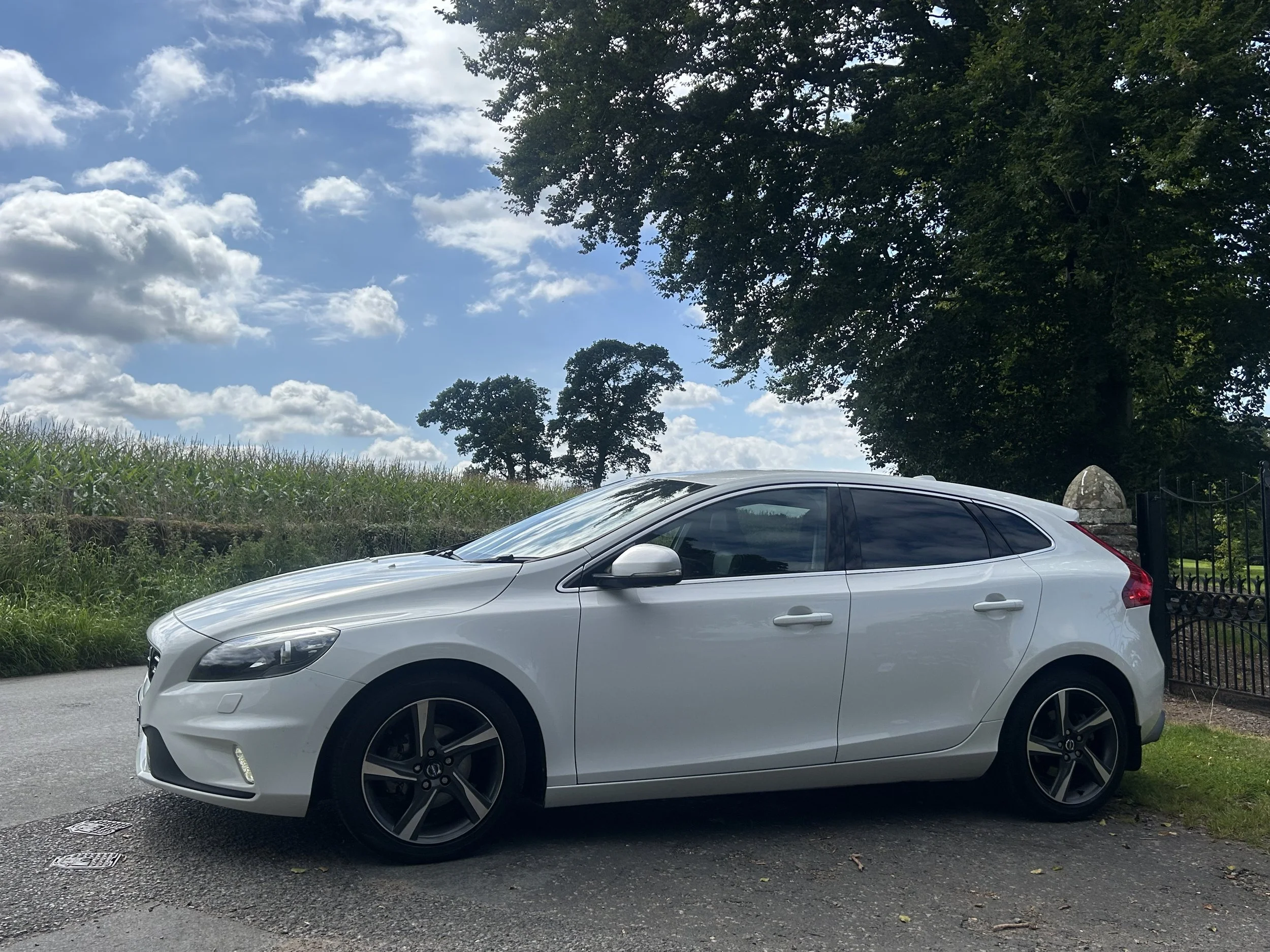 A white sedan car parked on a driveway next to green grass and trees under a partly cloudy sky.