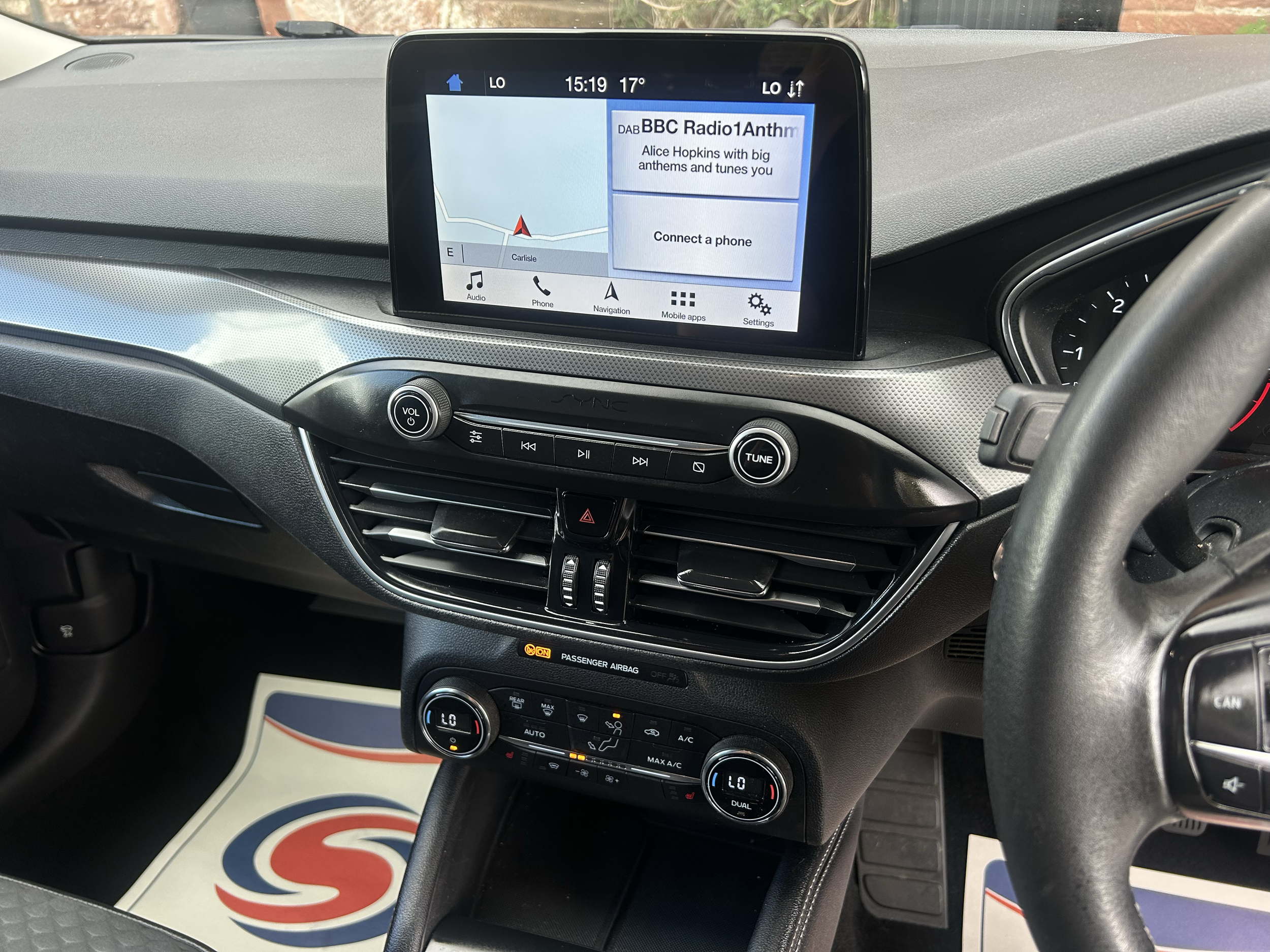 Car dashboard with a digital display showing radio station BBC Radio 1 Anthem, navigation map, and climate controls.