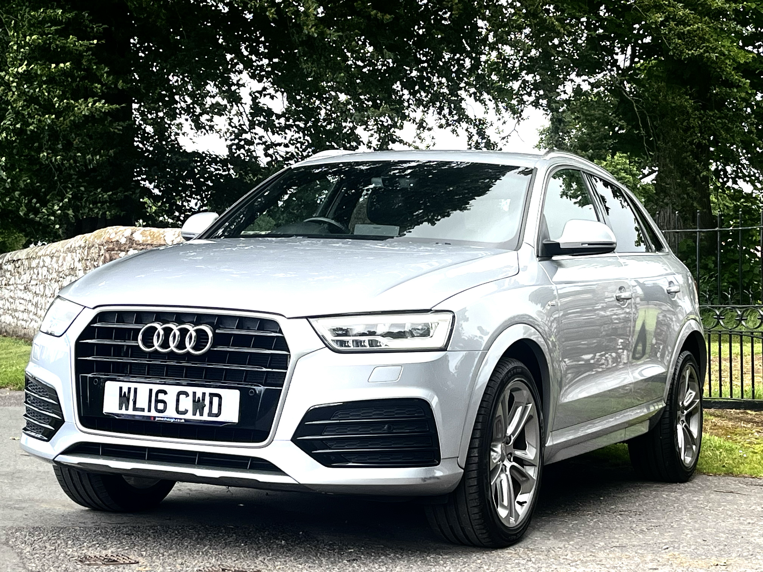 A silver Audi car parked on a street with trees and a stone wall in the background.