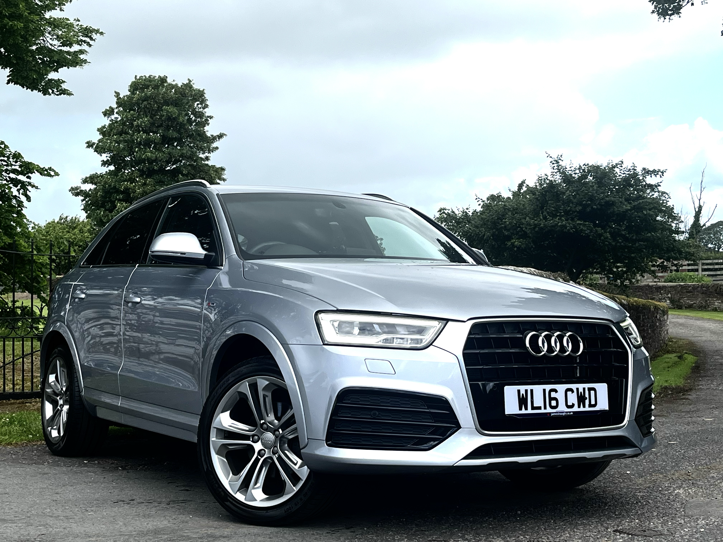 A silver Audi SUV parked on a paved surface next to a green grassy area with trees and a black metal fence in the background under a partly cloudy sky.