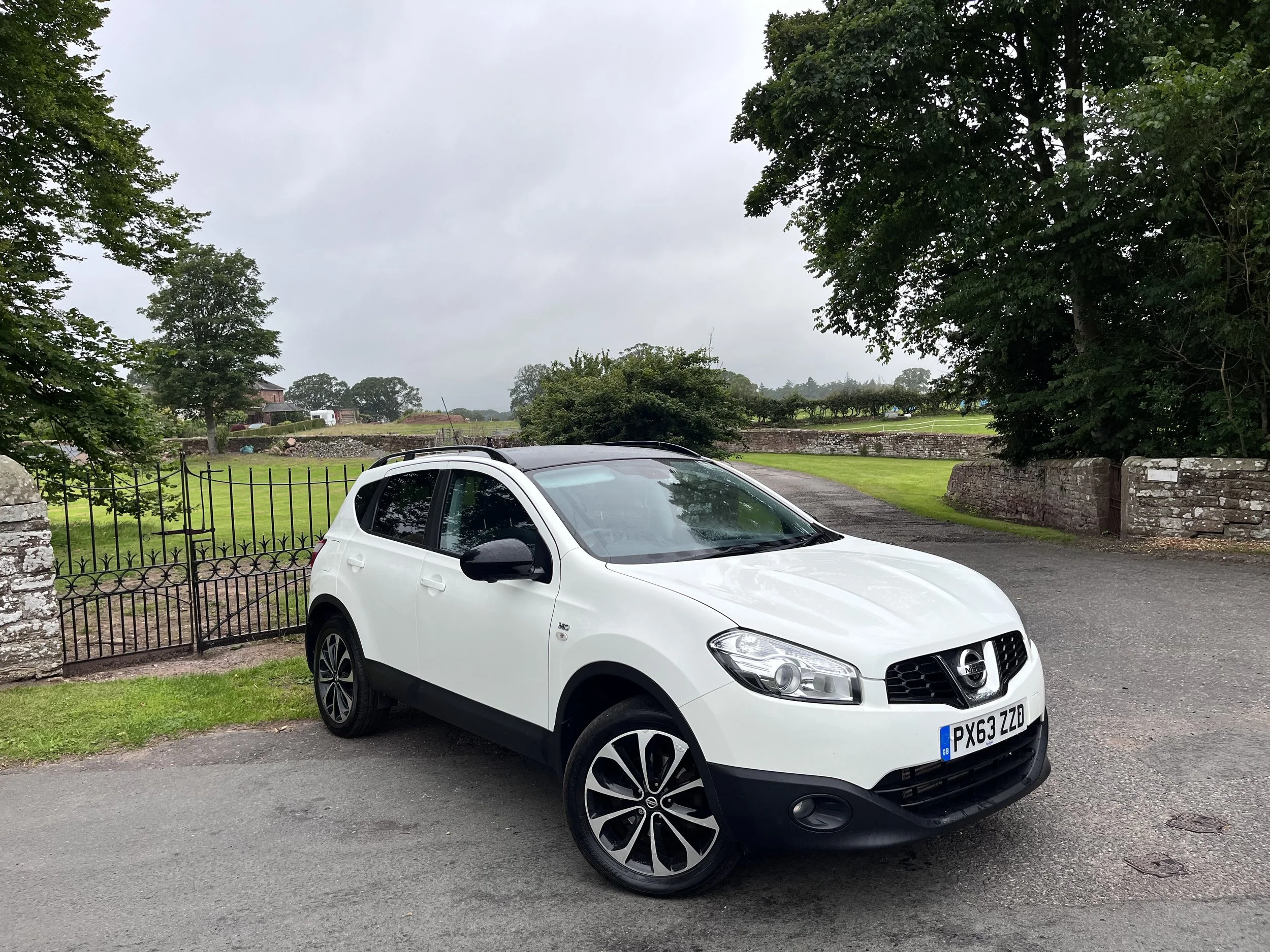 A white Nissan SUV parked on a paved driveway with green trees and a stone fence in the background.