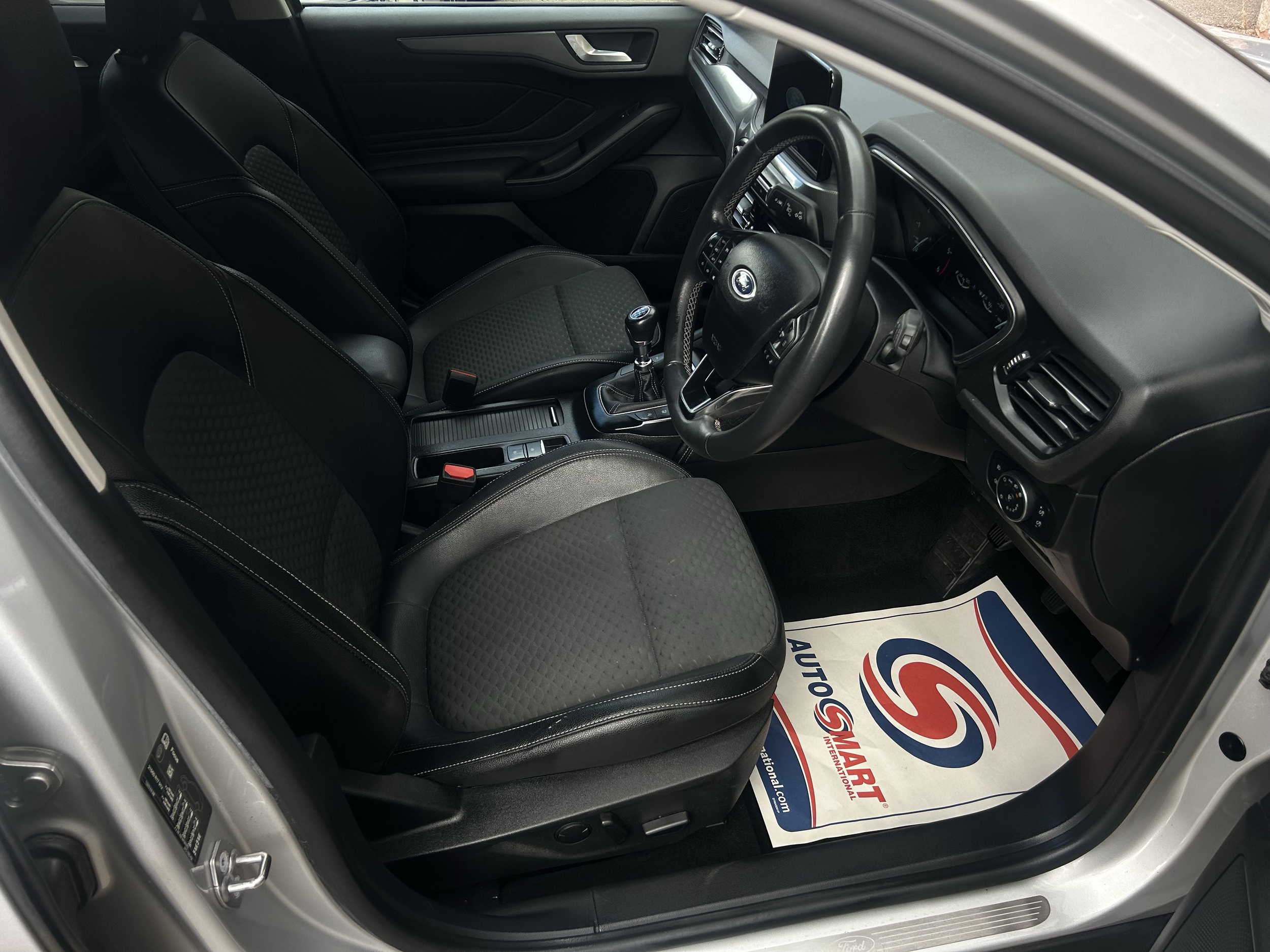 The interior of a modern car showing black leather seats, dashboard, steering wheel, and gear shift. A paper with a red, blue, and white logo, likely from a car dealership or service center, is on the floor.