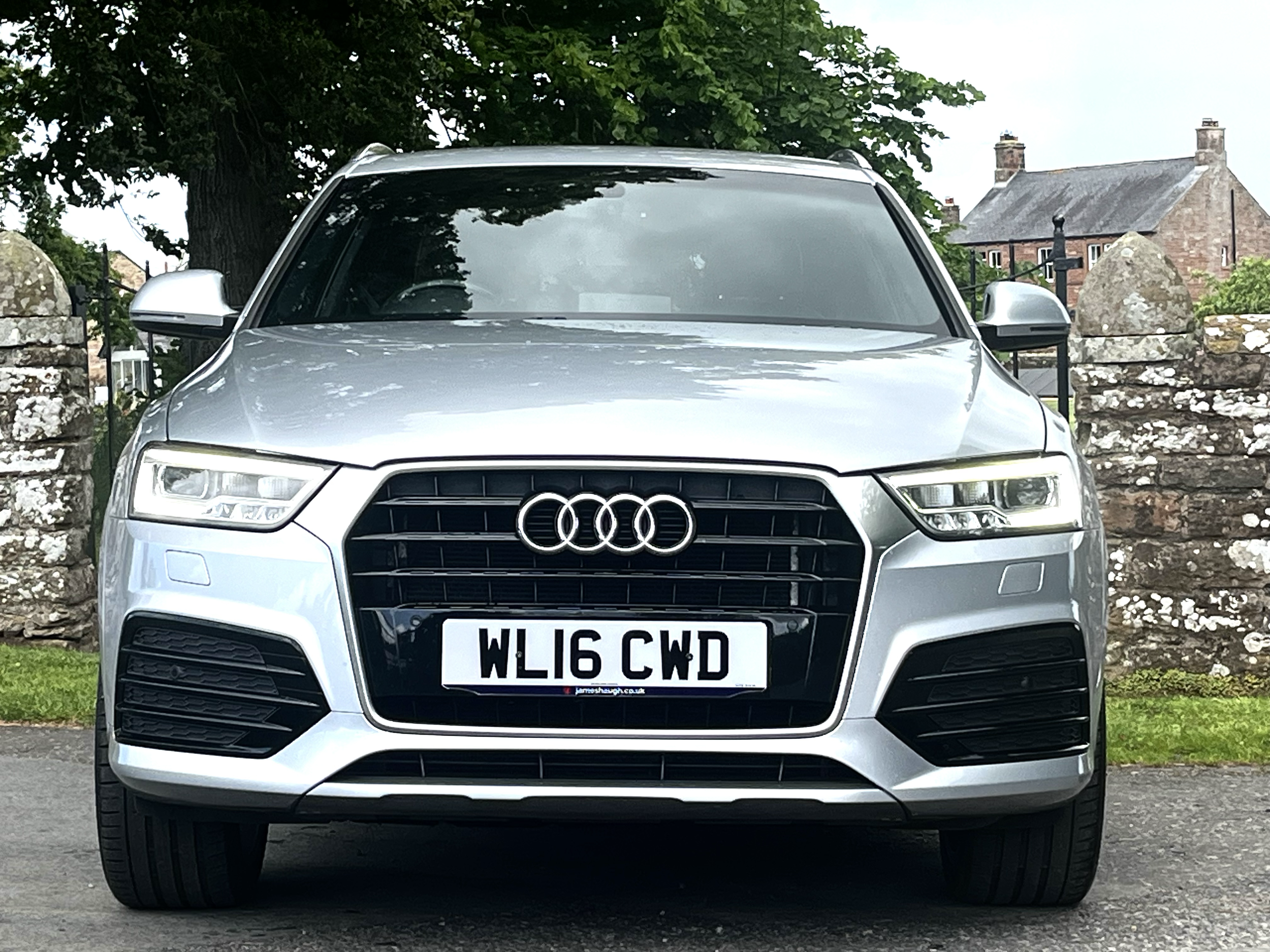 A silver Audi SUV parked in front of a stone fence with a rural house and trees in the background.
