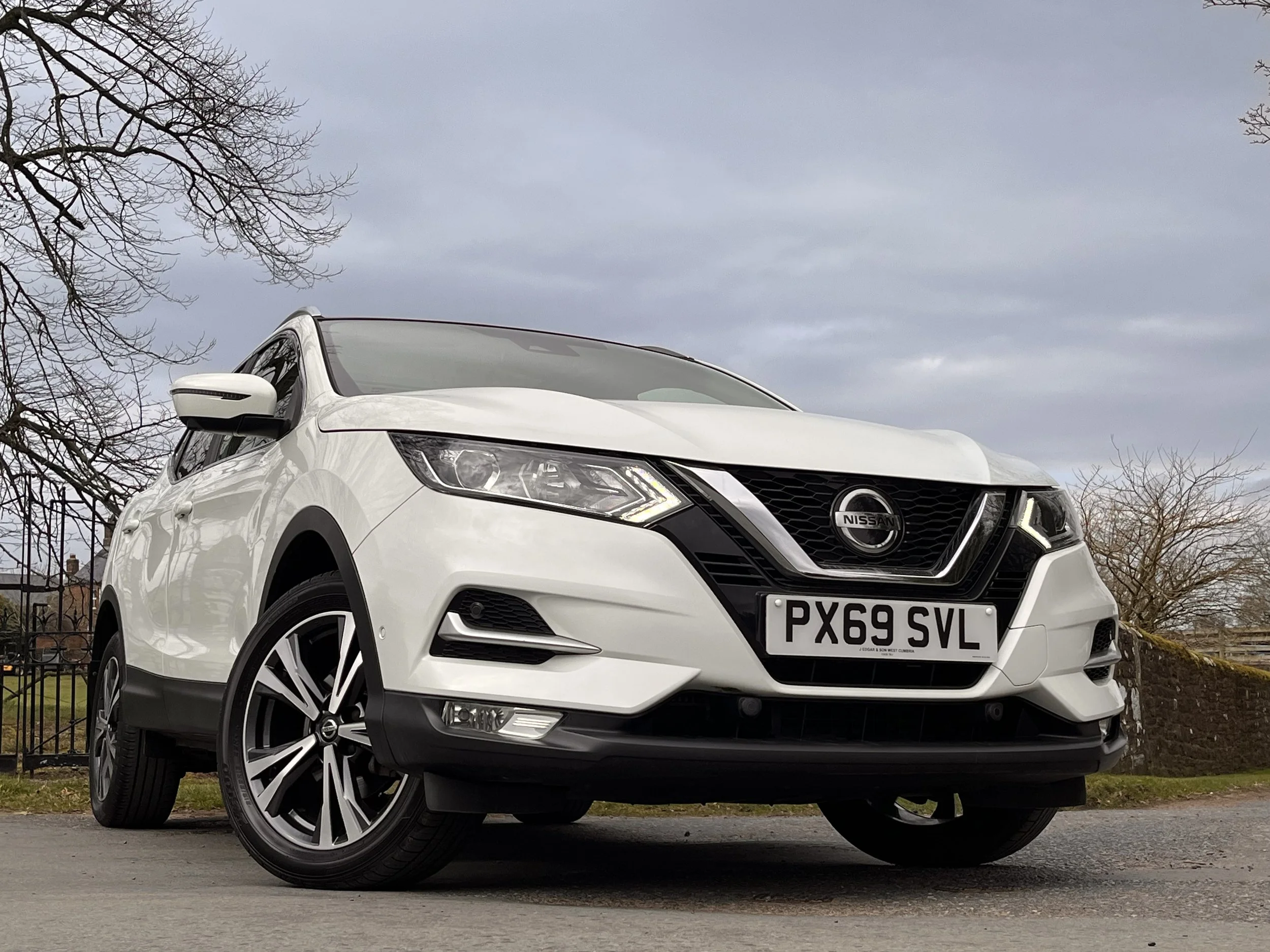 A white Nissan SUV parked outdoors on a paved surface with trees and a cloudy sky in the background.