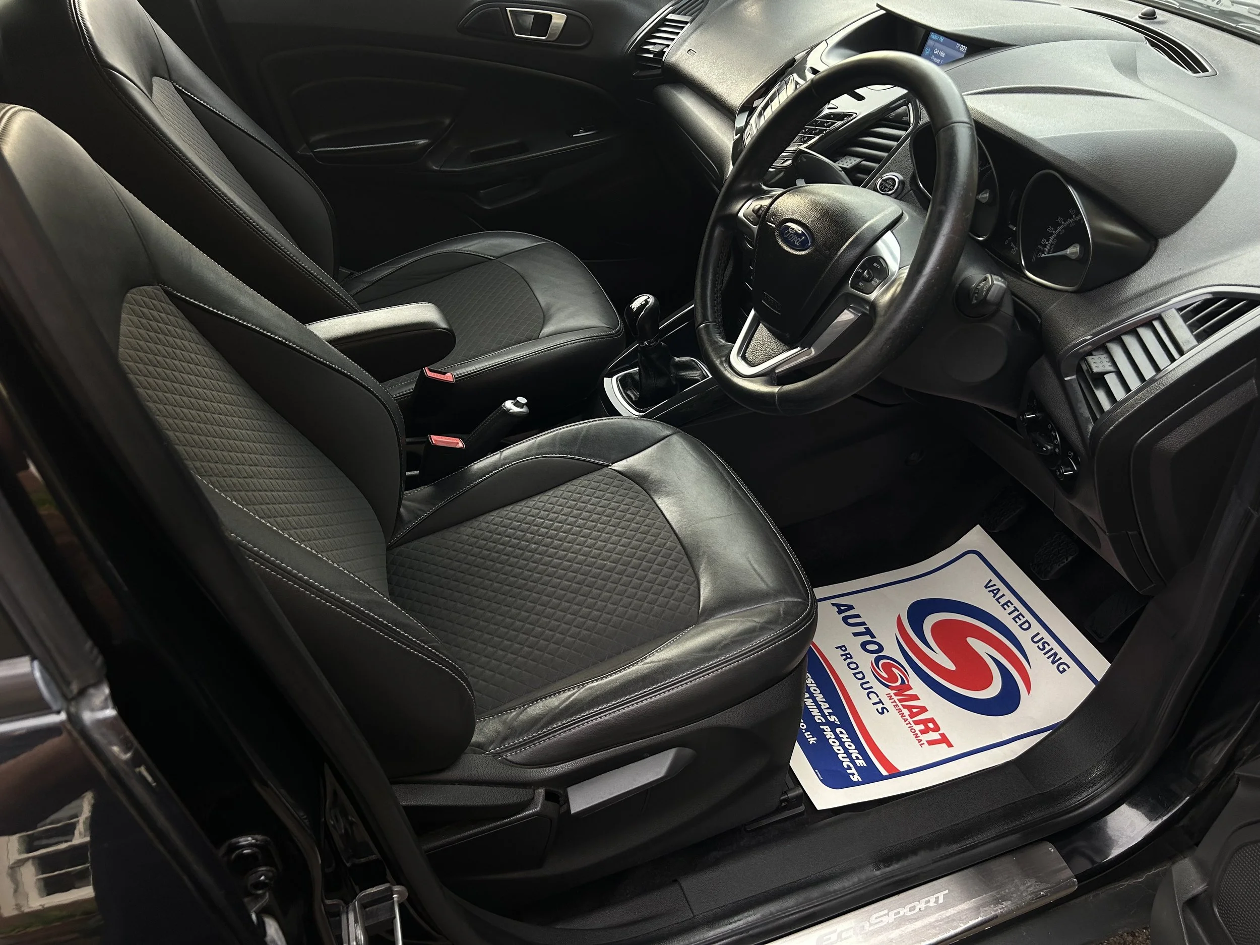 Interior view of a car showing black leather seats, steering wheel, dashboard, and a floor mat with a logo and text for 'AutoSmart' and 'Valeted Using'.