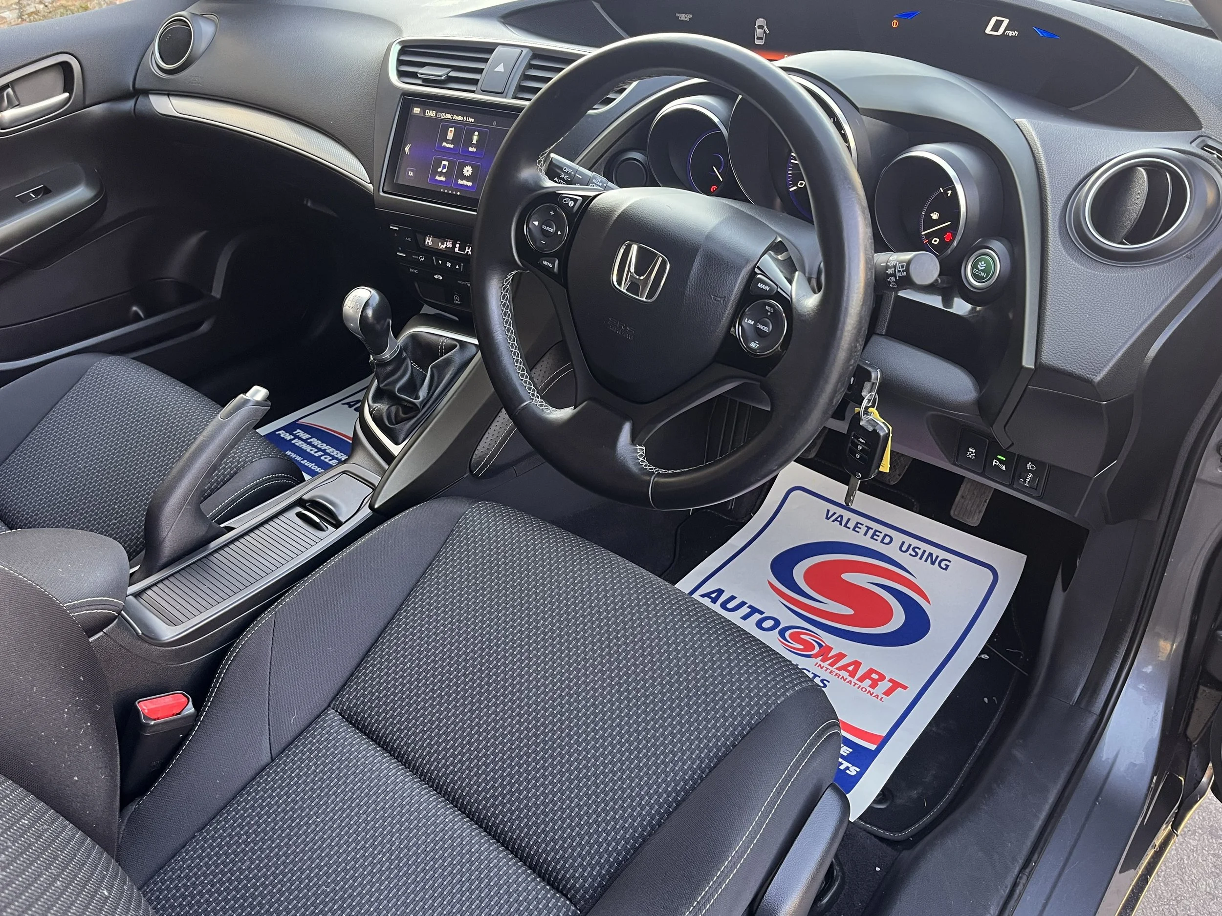 Interior of a car showing the dashboard, steering wheel, gear shift, and front seats, with a 'Valeted Using AutoSmart' floor mat.