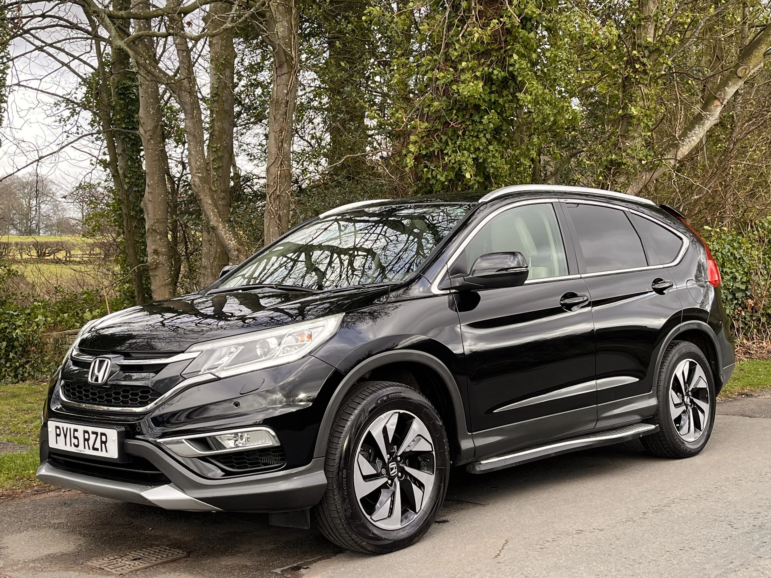 A black Honda HR-V parked on a paved street with trees and bushes in the background.