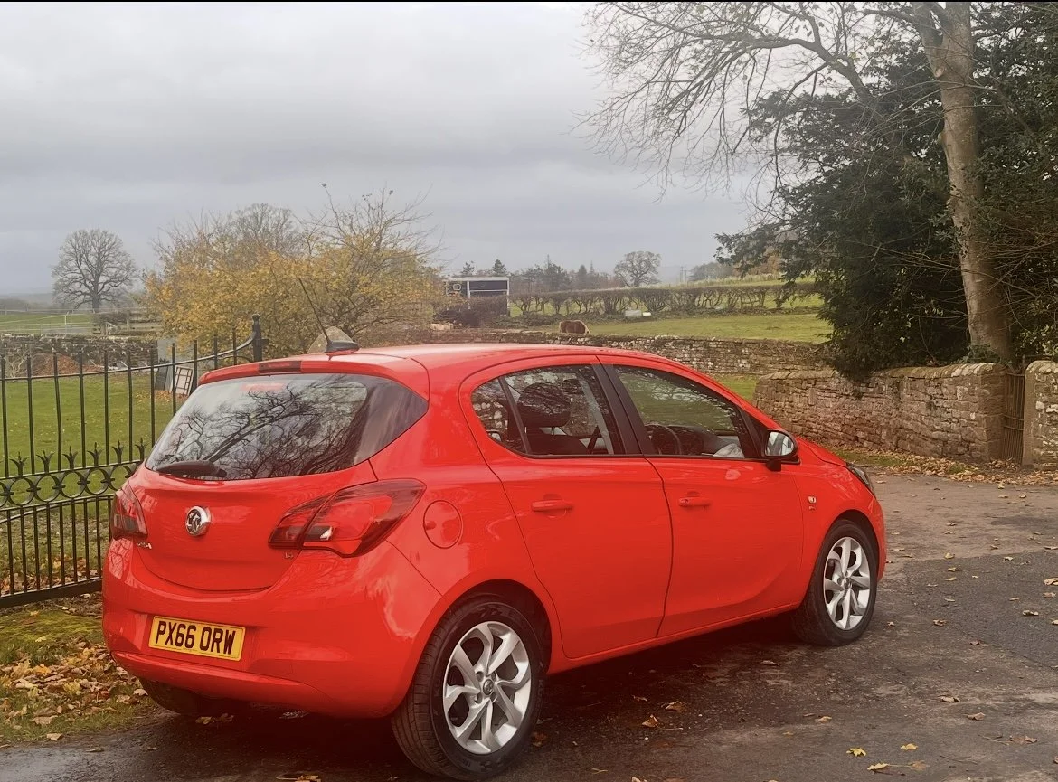 Red hatchback car parked on a paved area near a stone wall and metal fence with autumn trees and grassy fields in the background under a cloudy sky.