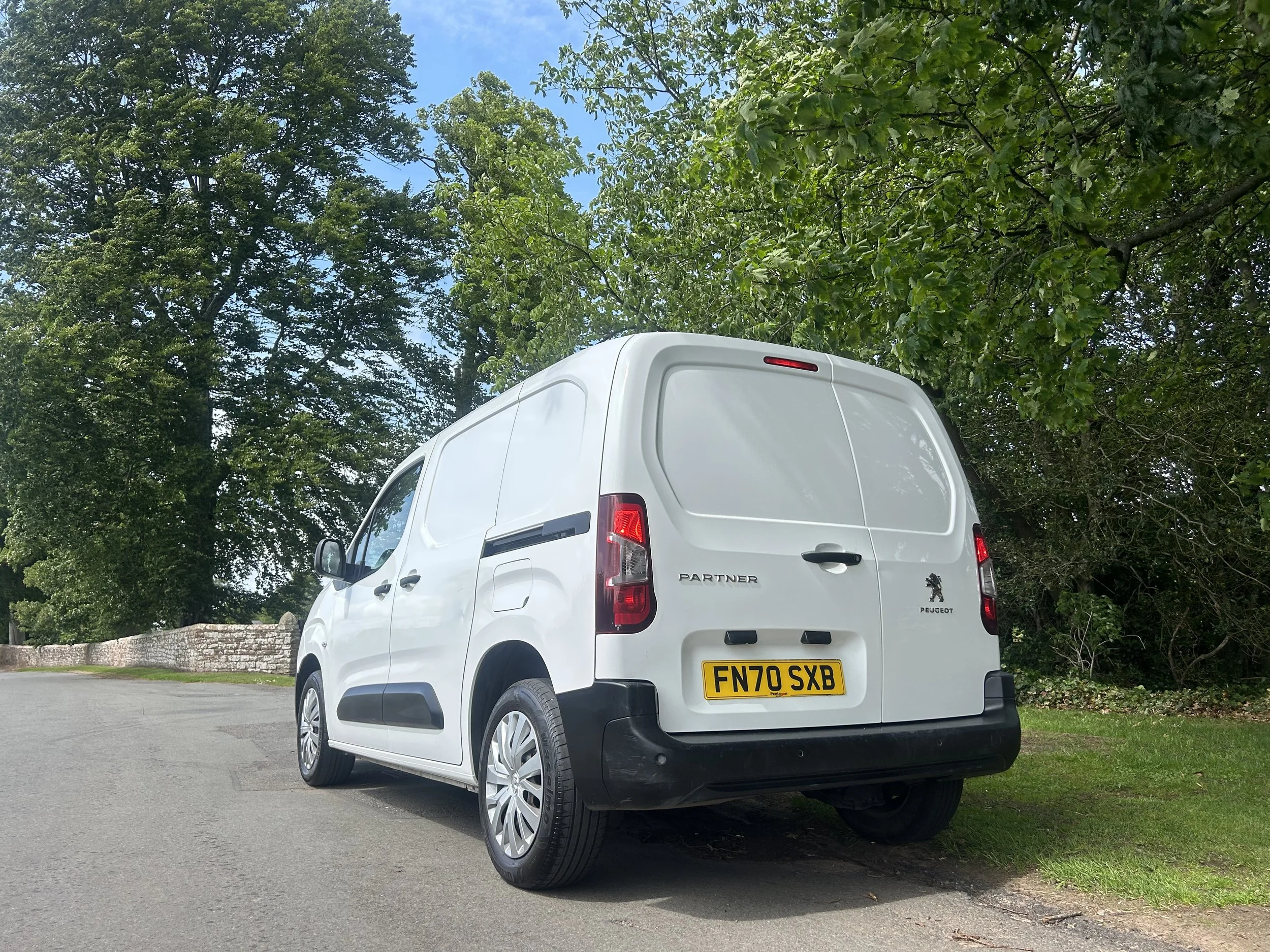 White Peugeot Partner van parked near a grassy area with trees and a stone wall in the background.