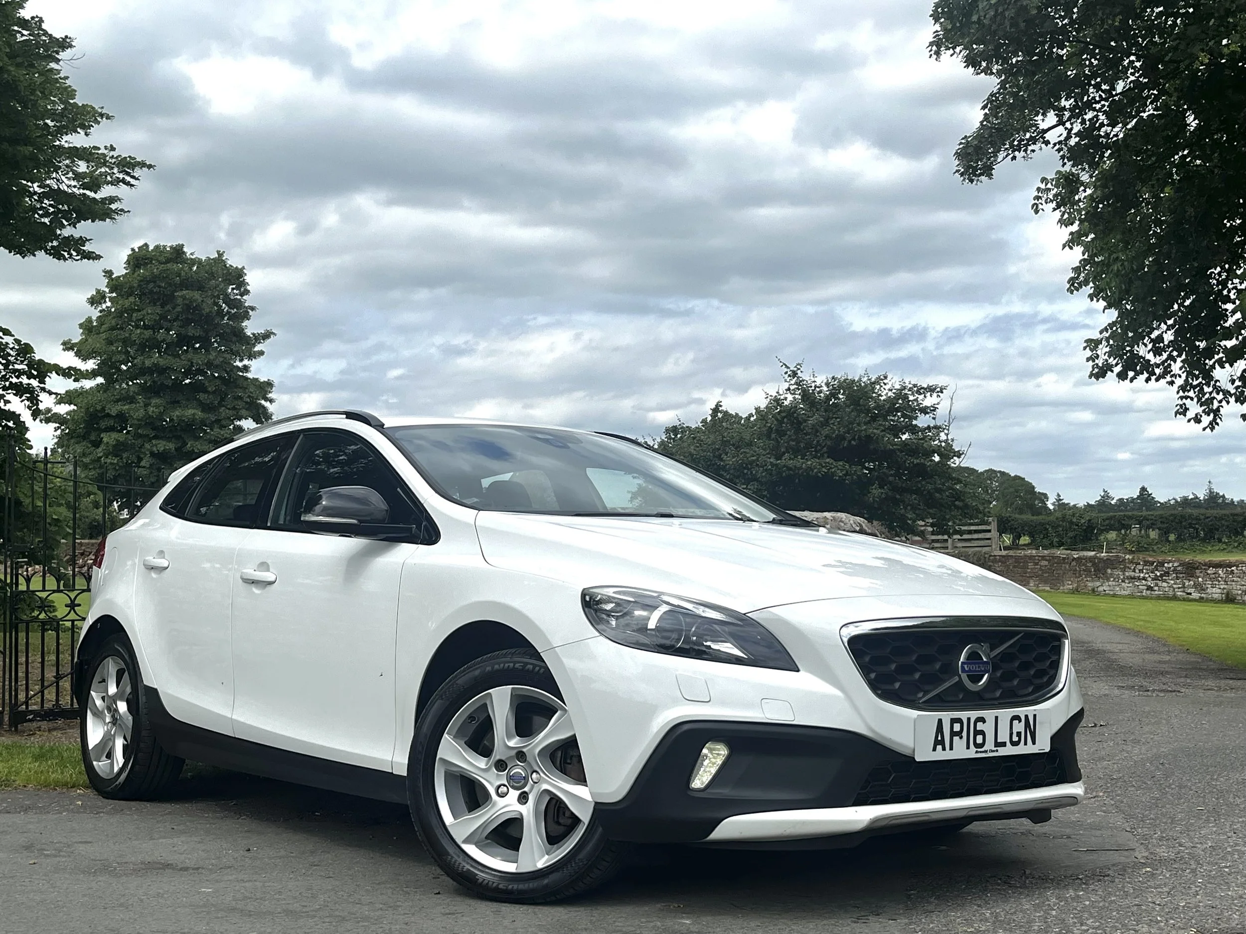 A white Volvo car parked outdoors on a cloudy day with trees and a fence in the background.