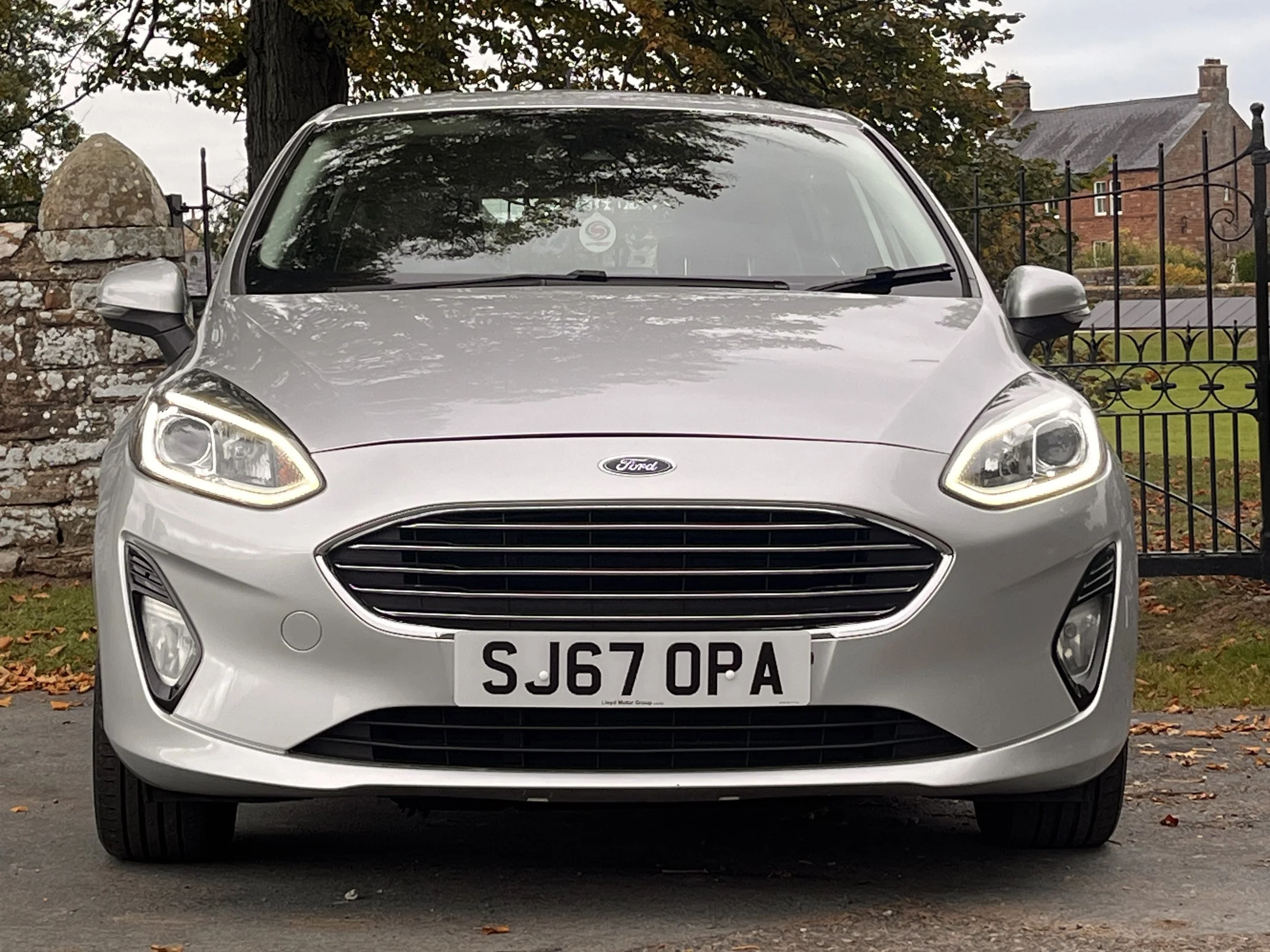 Front view of a silver Ford Focus parked outdoors with a brick wall, tree, and buildings in the background.