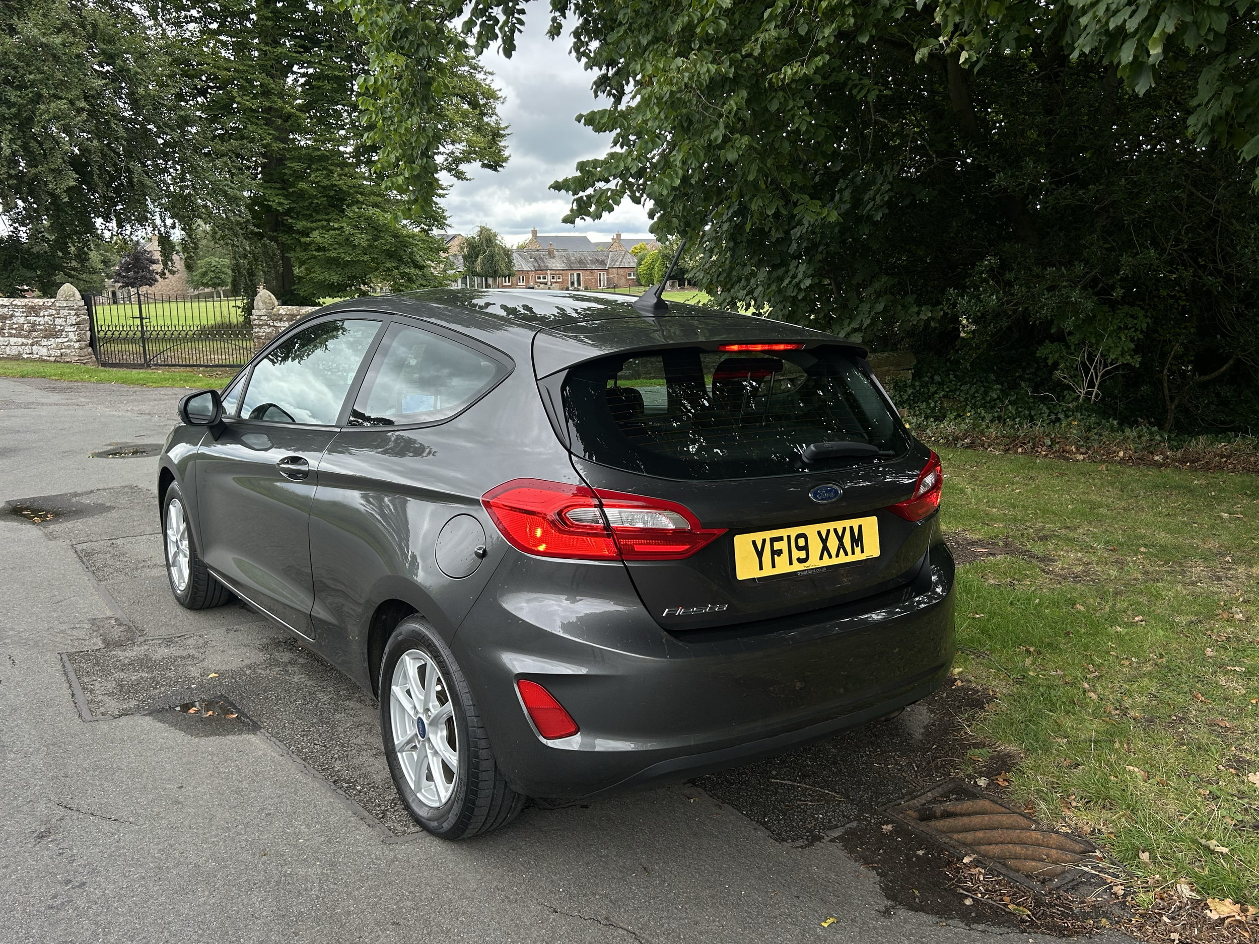 A gray Ford Fiesta parked on a street next to a green grassy area with trees and a brick wall with an iron gate. There are trees and houses in the background.