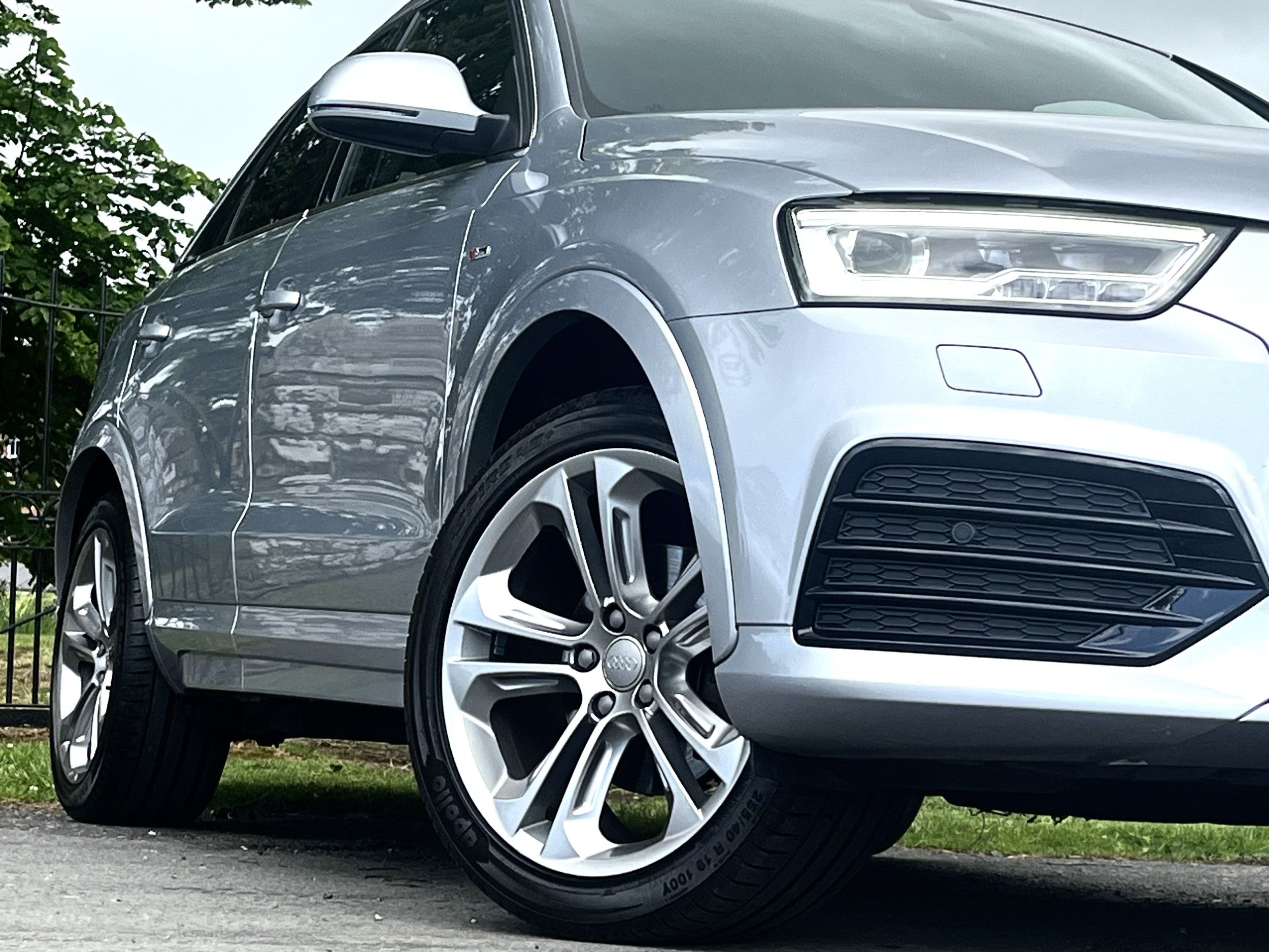 Close-up of a silver Audi car parked outdoors, with visible front wheel, headlights, side mirror, and part of the front grille. Green trees and a fence are in the background.