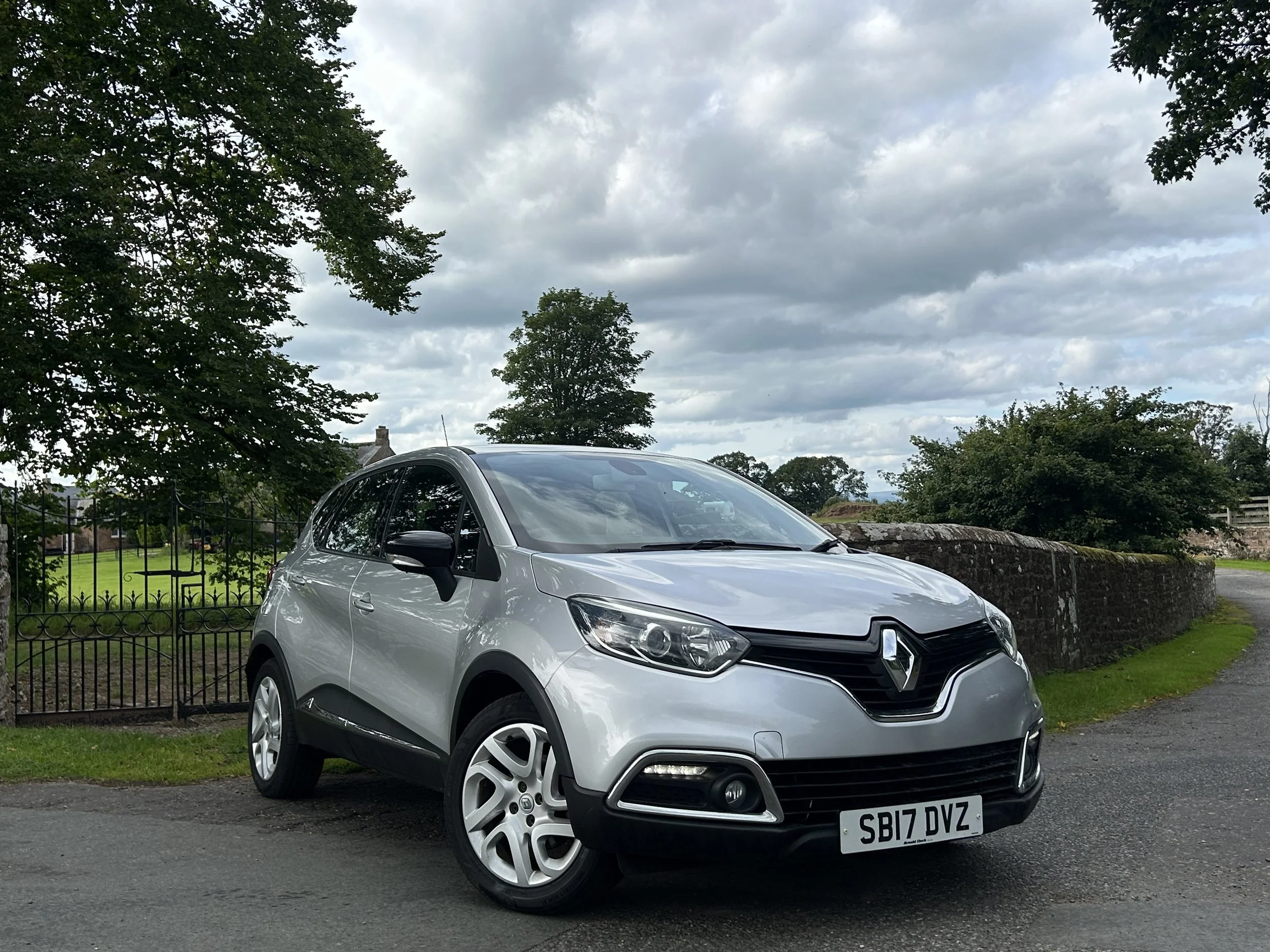 A silver Renault car parked on a road with grassy area and trees in the background under a cloudy sky.