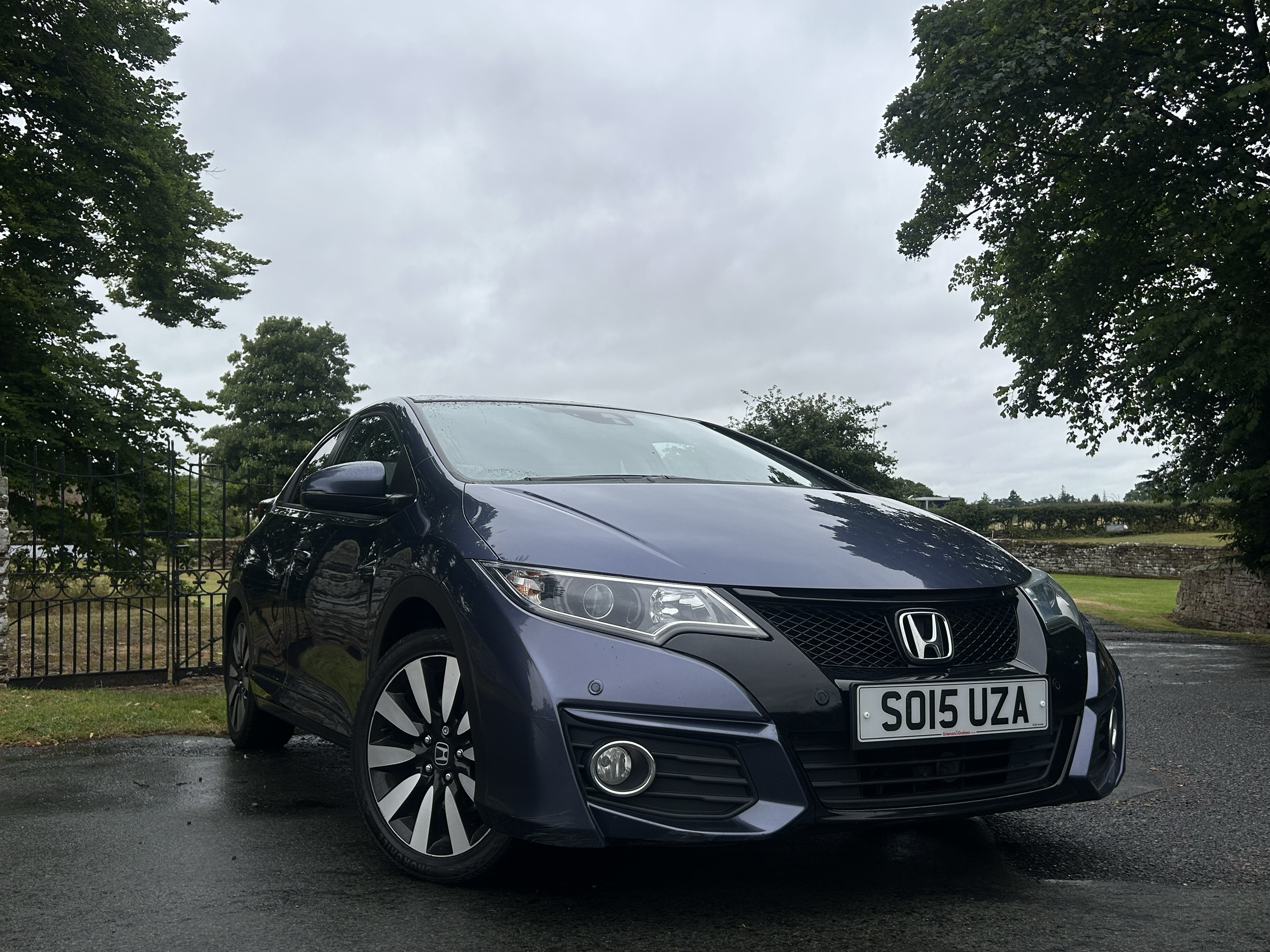 A black Honda car parked on the street with greenery and trees in the background, and an overcast sky above.