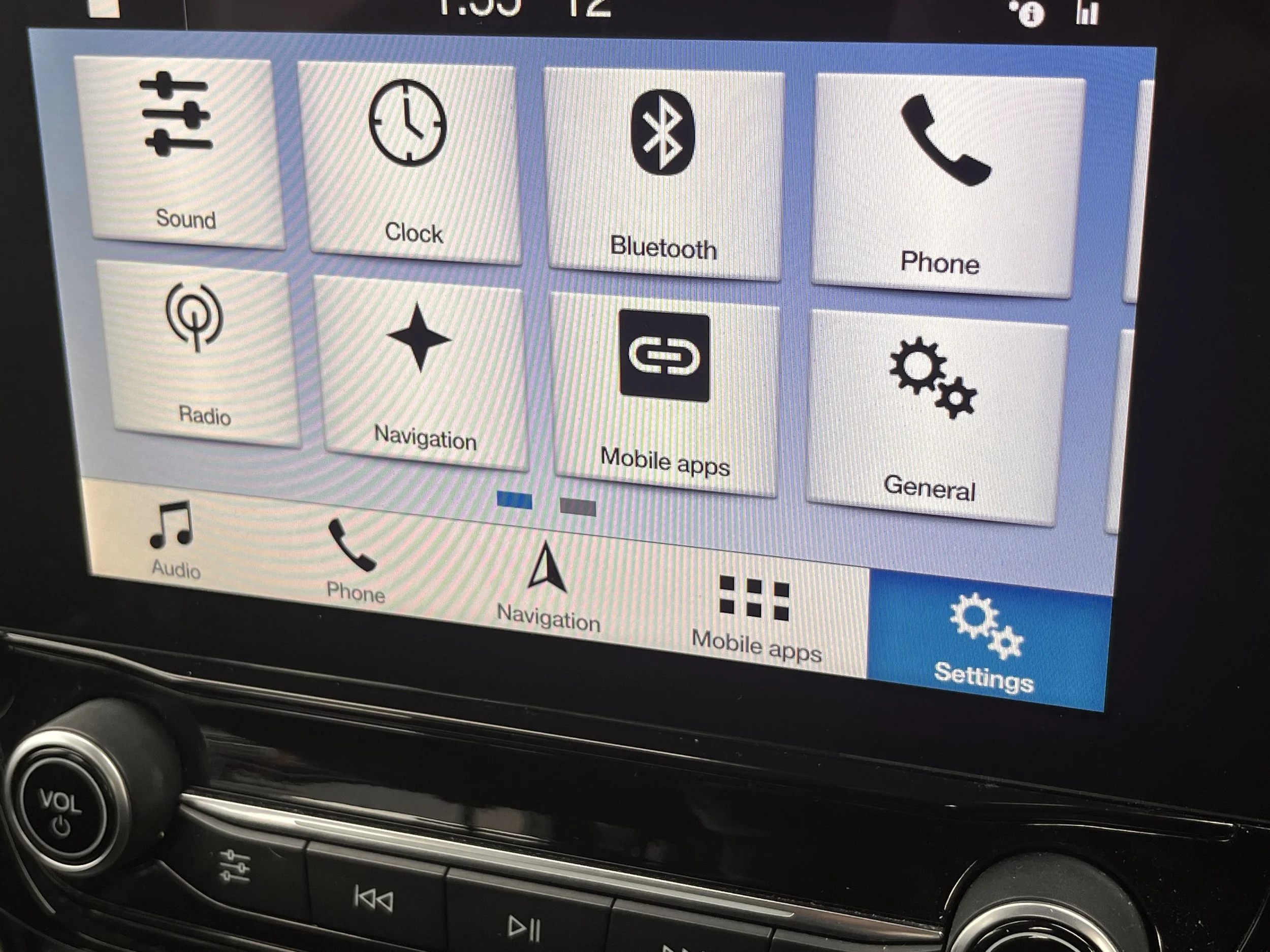 Car infotainment system screen displaying various icons including Sound, Clock, Bluetooth, Phone, Radio, Navigation, Mobile Apps, and General. There are additional options for Audio, Phone, Navigation, Mobile Apps, and Settings in a toolbar at the bo