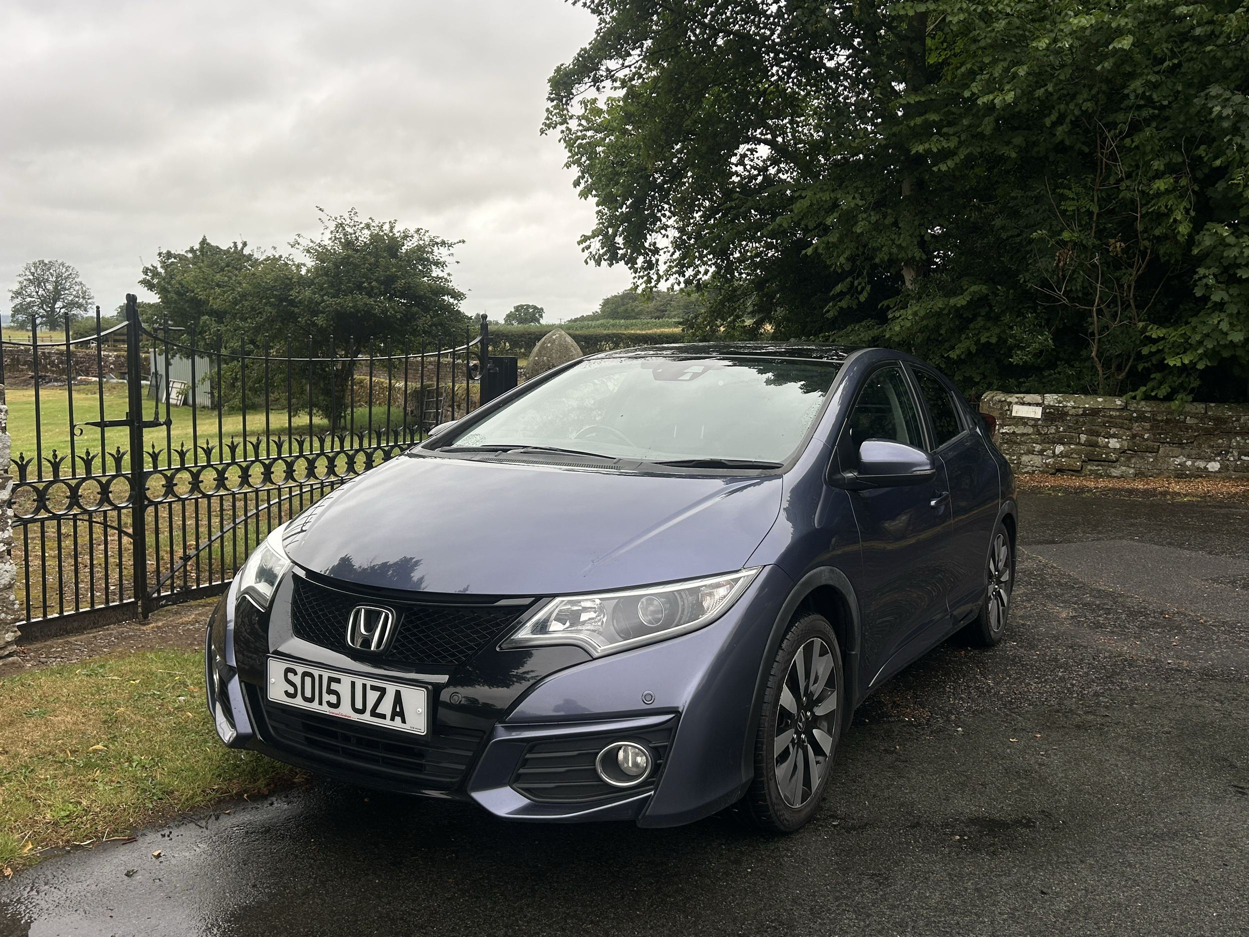 A gray Honda Civic parked on a wet asphalt surface in front of a black metal fence, with green trees and cloudy sky in the background.