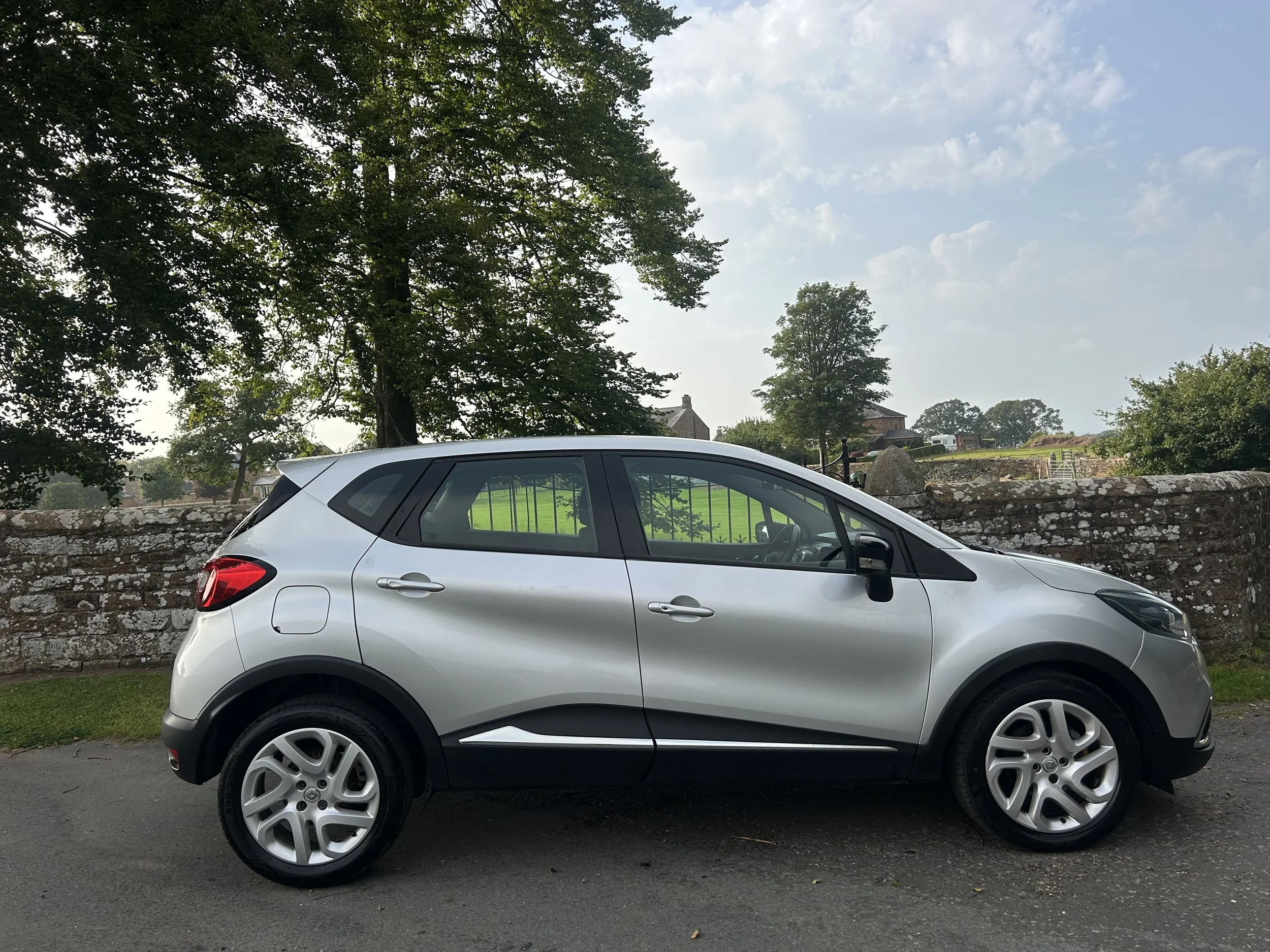 Silver compact SUV parked on a street with a stone wall and green trees in the background.