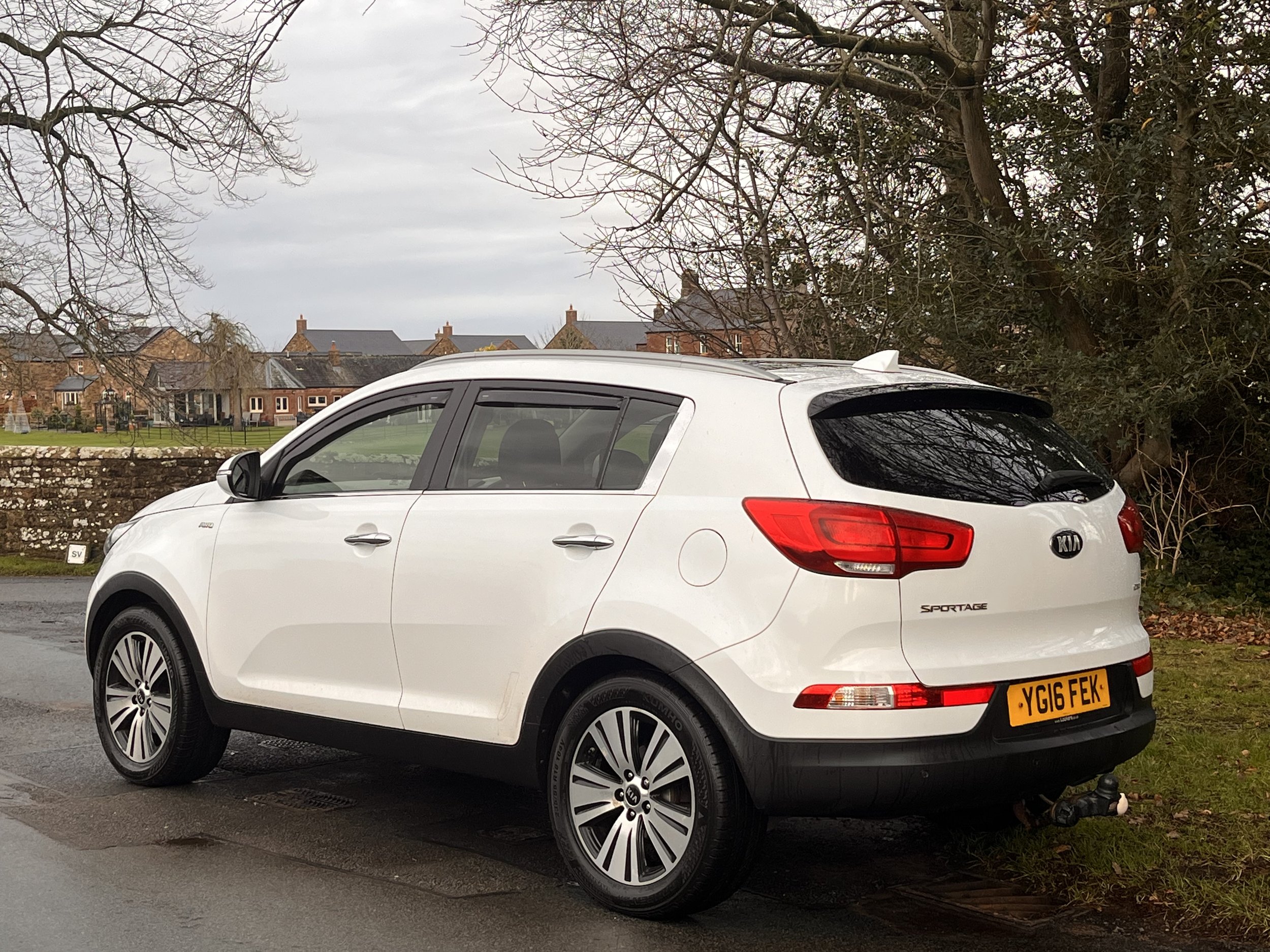 A white Kia Sportage parked on the side of a road next to a grassy area with trees and houses in the background. It's a cloudy day.
