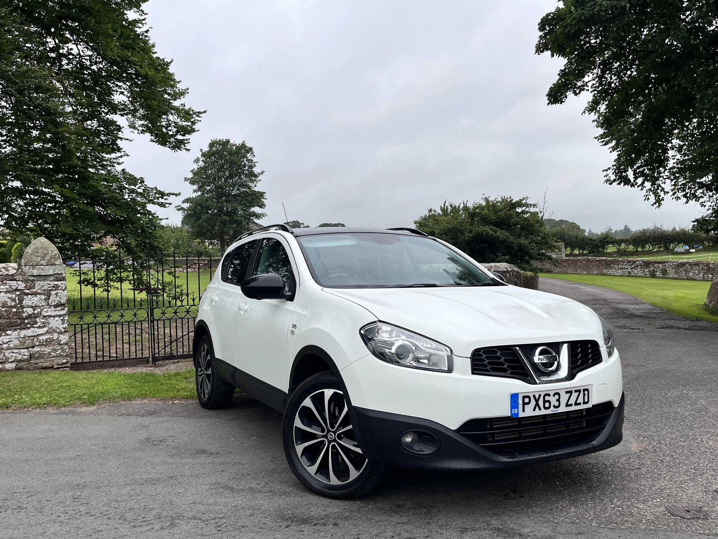 White Nissan compact SUV parked on a pavement with a green grassy area, trees, and cloudy sky in the background.