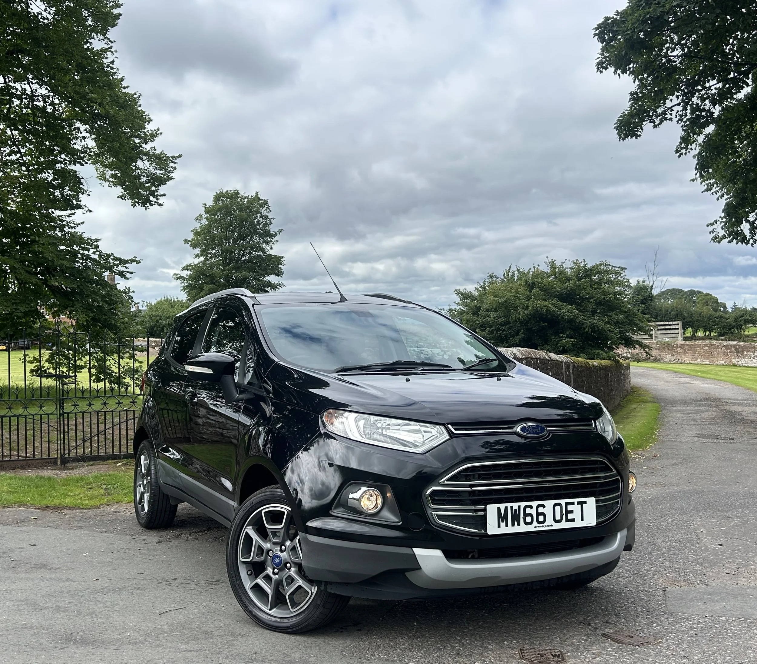 Black Ford EcoSport parked on a gravel driveway with trees, a stone wall, and cloudy sky in the background.