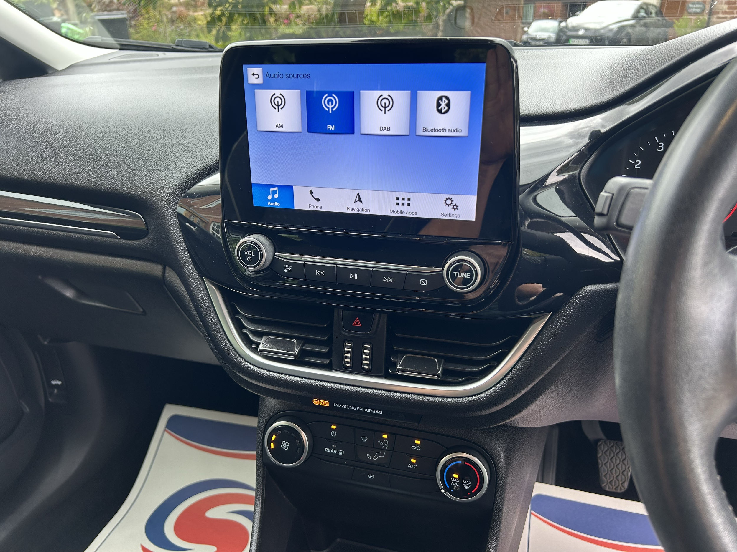The image shows a car's dashboard with a touchscreen displaying audio sources like AM, FM, DAB, and Bluetooth audio. There are also climate control buttons below the screen and a passenger airbag indicator. Outside the windshield, a parking lot with 