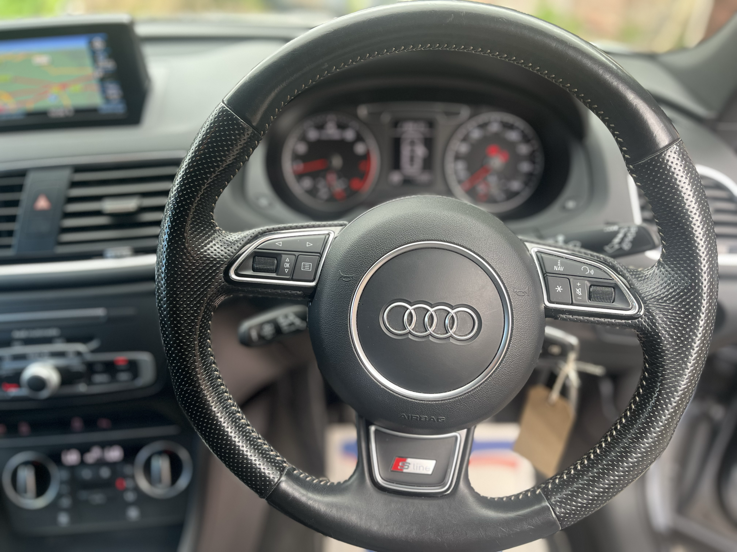 Interior view of a car dashboard with a black leather steering wheel featuring an Audi logo in the center and control buttons on both sides.