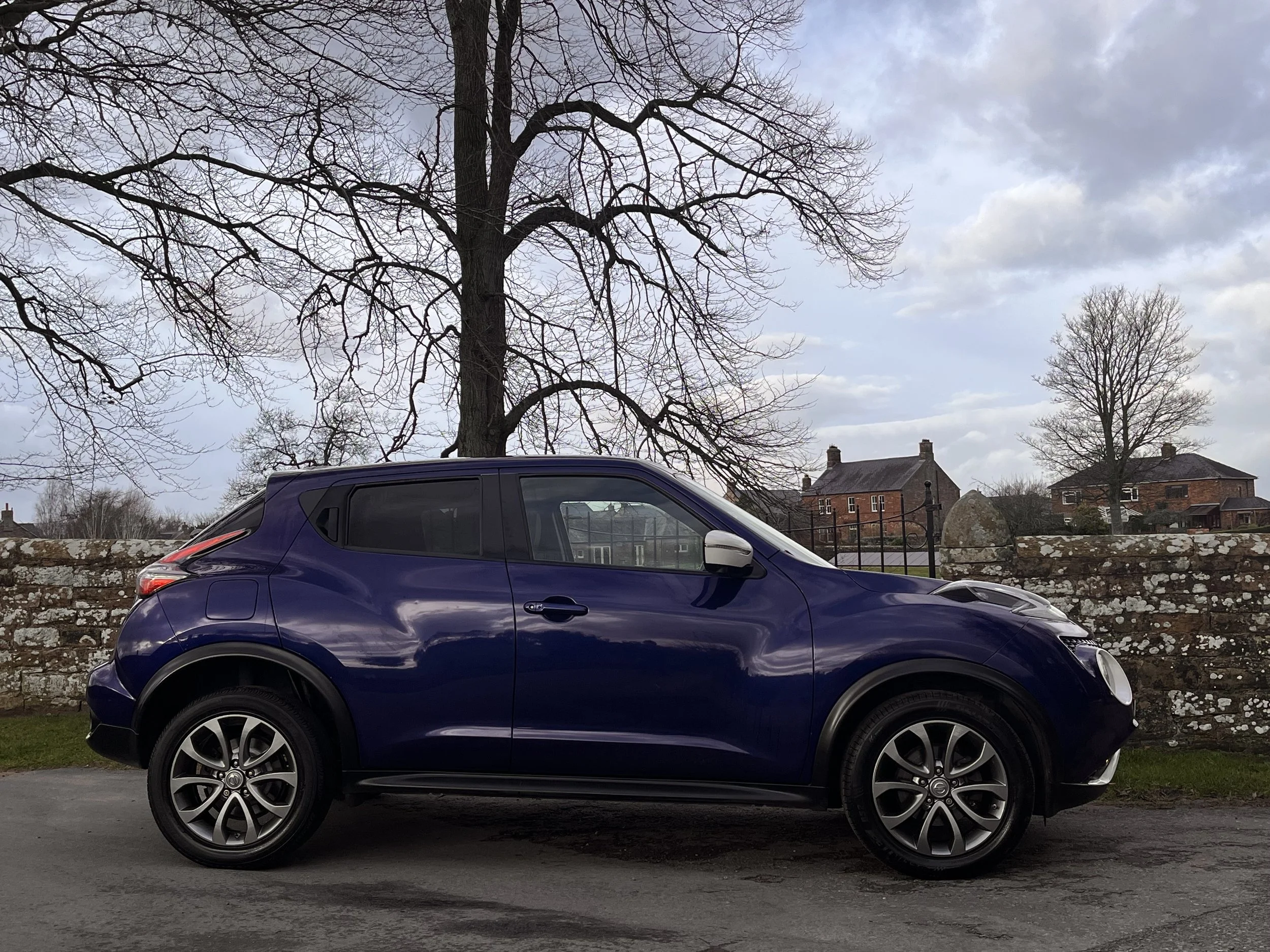 A blue compact crossover SUV parked outdoors on a paved surface with a stone wall, leafless trees, and houses in the background under a partly cloudy sky.