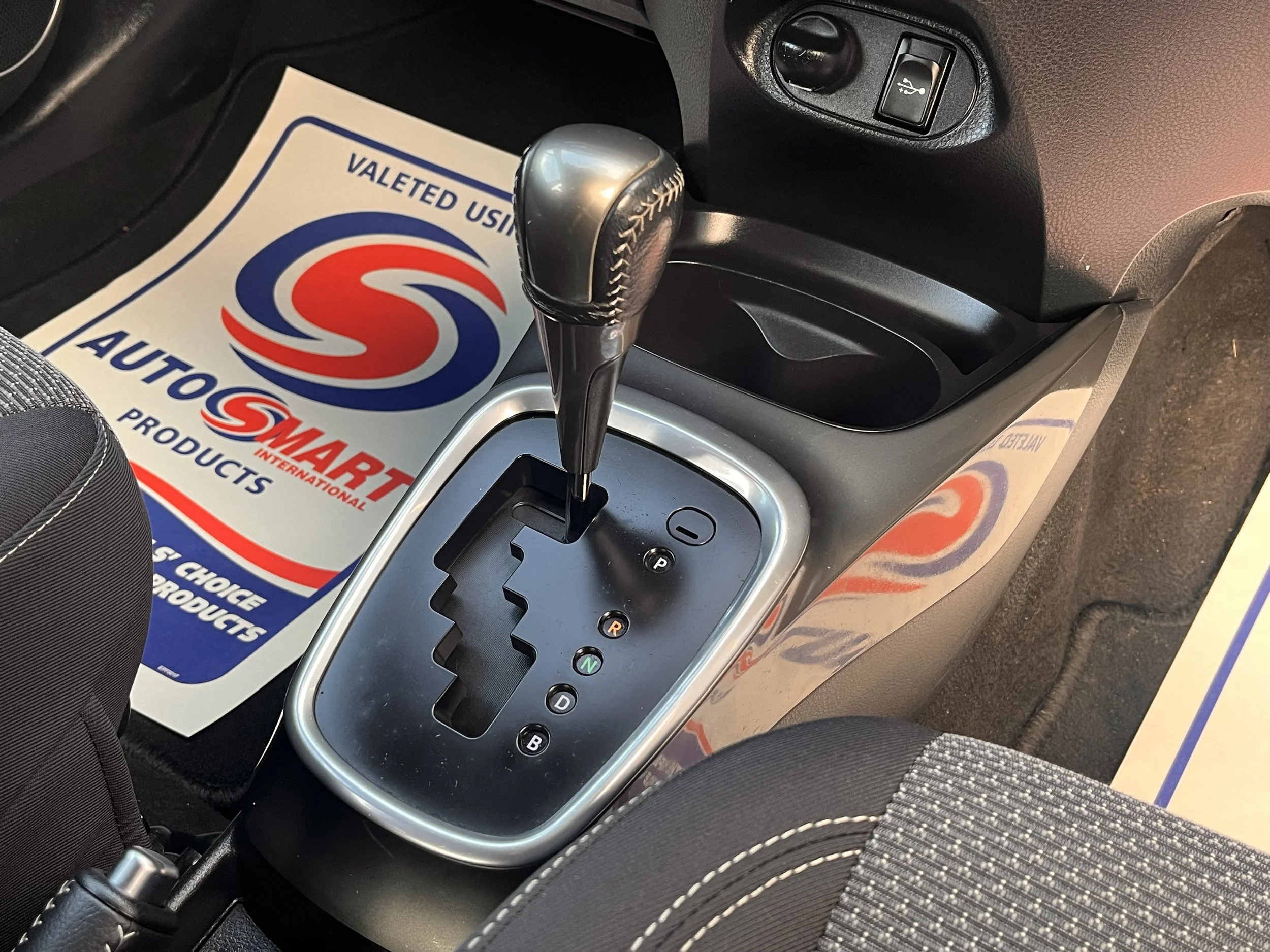 The image shows a car's automatic gear shift lever in park, located in the center console, with a promotional paper mat for AutoSmart Products on the floor.