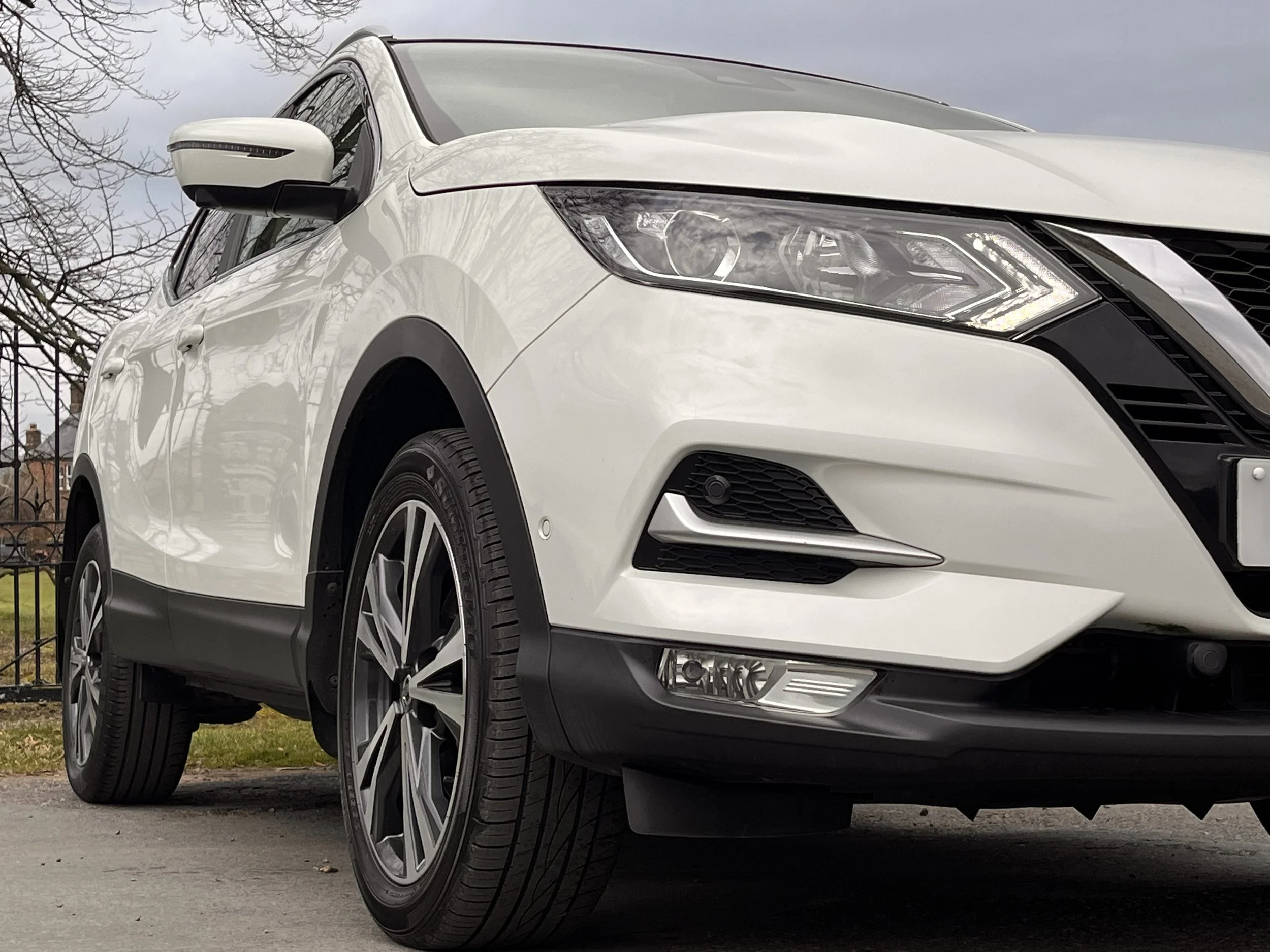 Front-left view of a white SUV with modern headlights, black grille, and alloy wheels parked on a driveway with trees and a cloudy sky in the background.