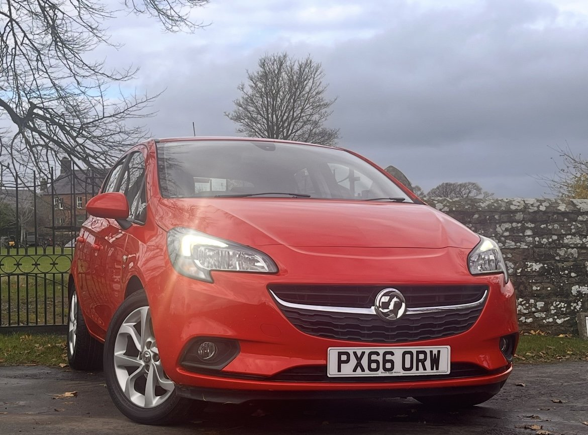 A red Vauxhall Corsa hatchback car parked outdoors on a paved area with a stone wall, green grass, and trees in the background under a cloudy sky.