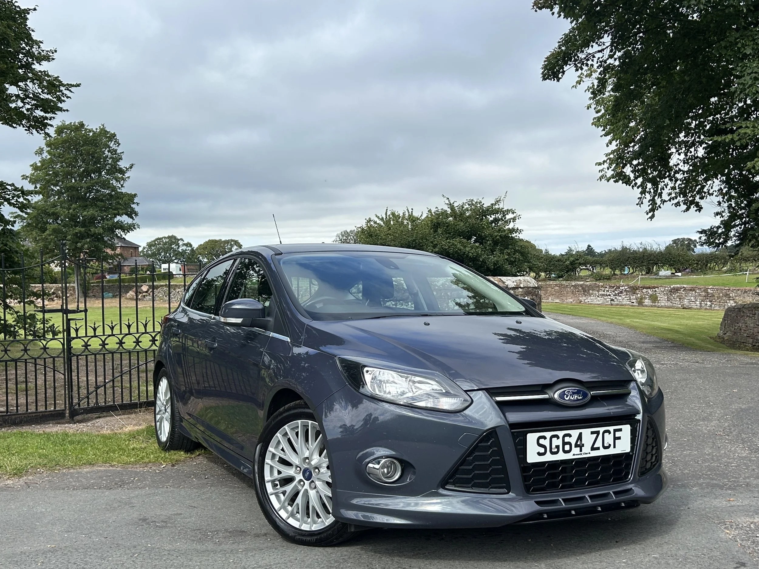 A dark gray Ford Fiesta hatchback parked on a paved surface near a grassy area with trees and a stone wall in the background.
