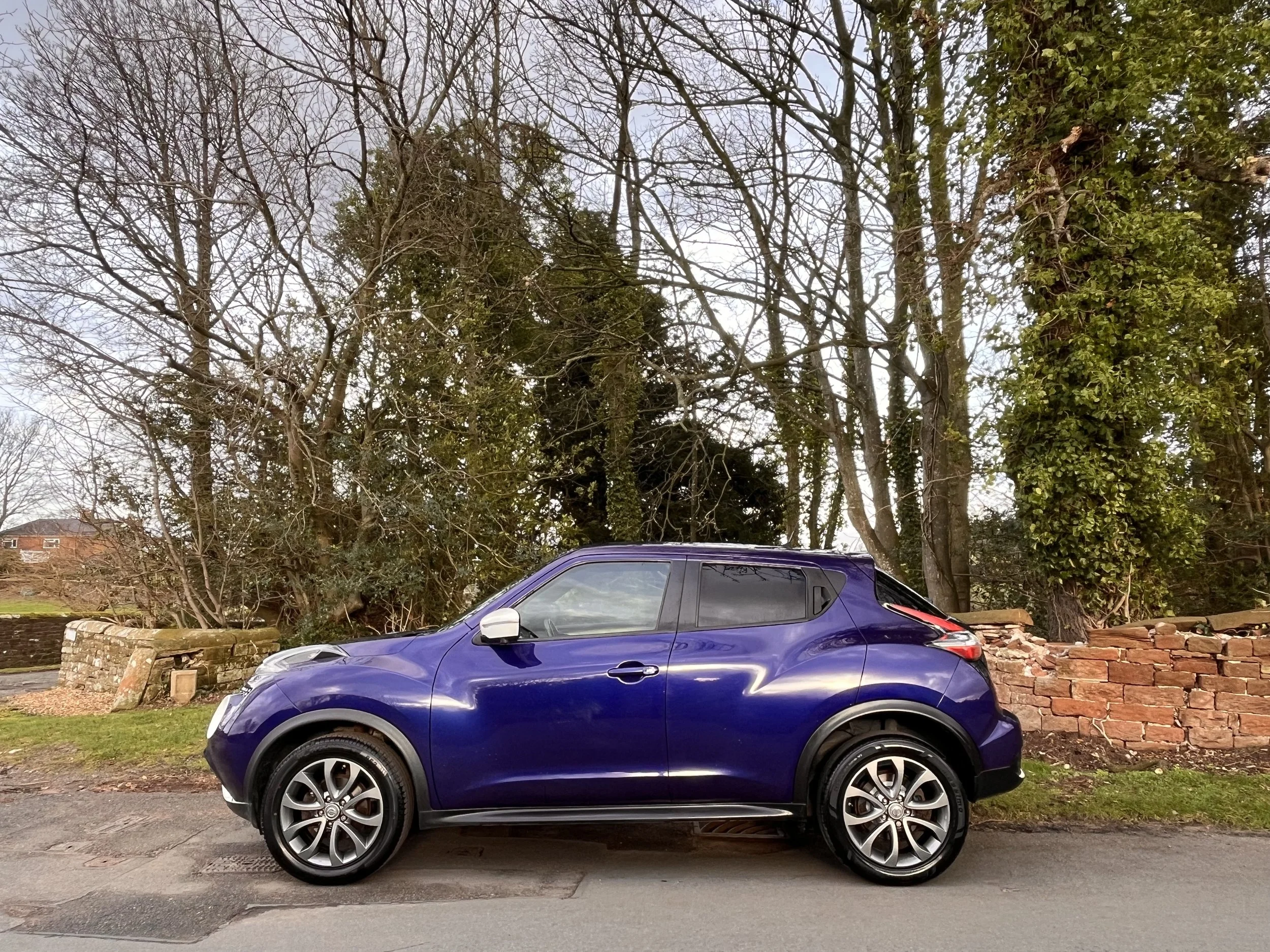 A blue compact car parked on the side of a street in front of a brick wall and trees.