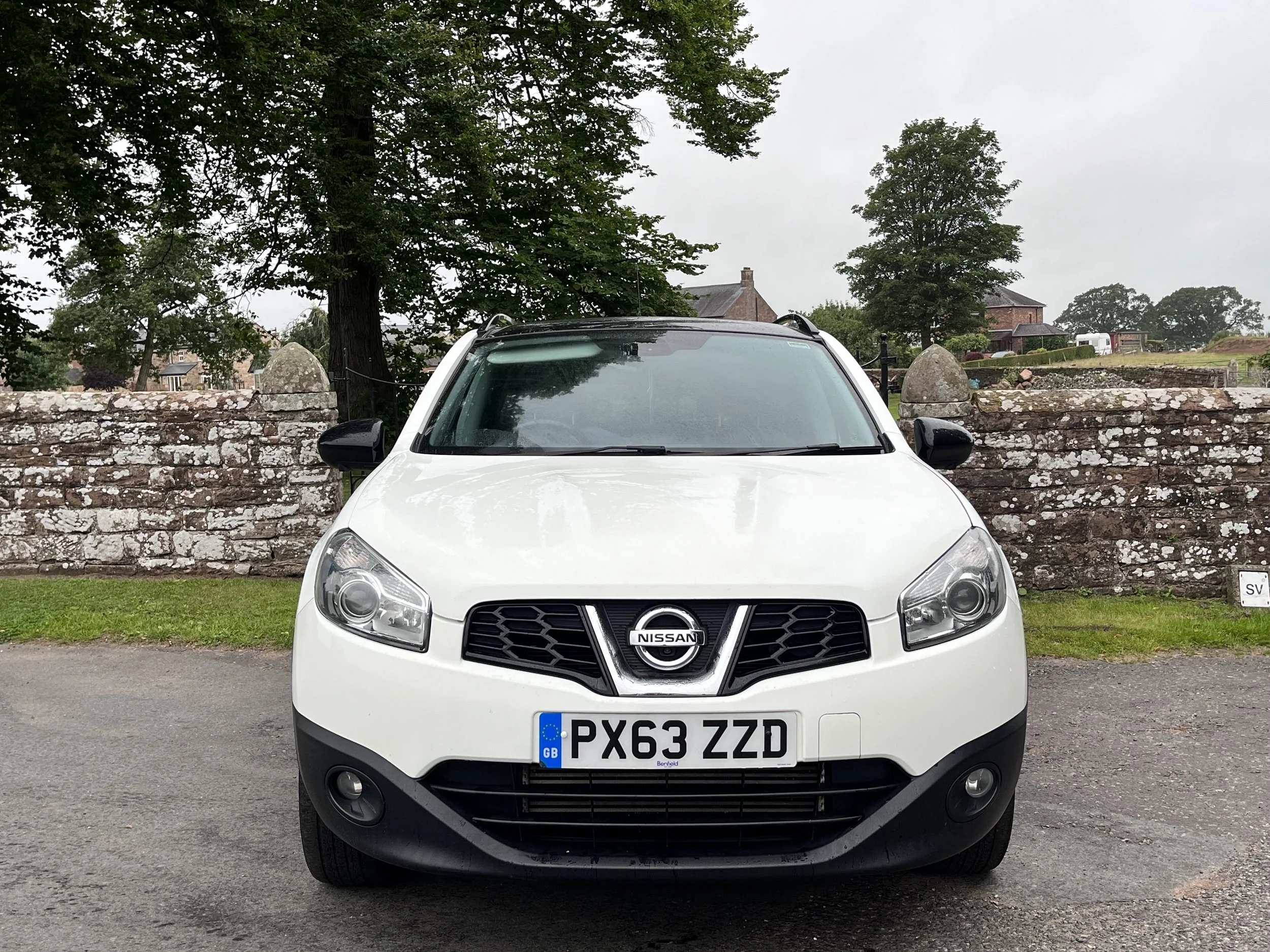 Front view of a white Nissan SUV parked on a paved surface in front of a stone wall, with trees and buildings in the background under a cloudy sky.