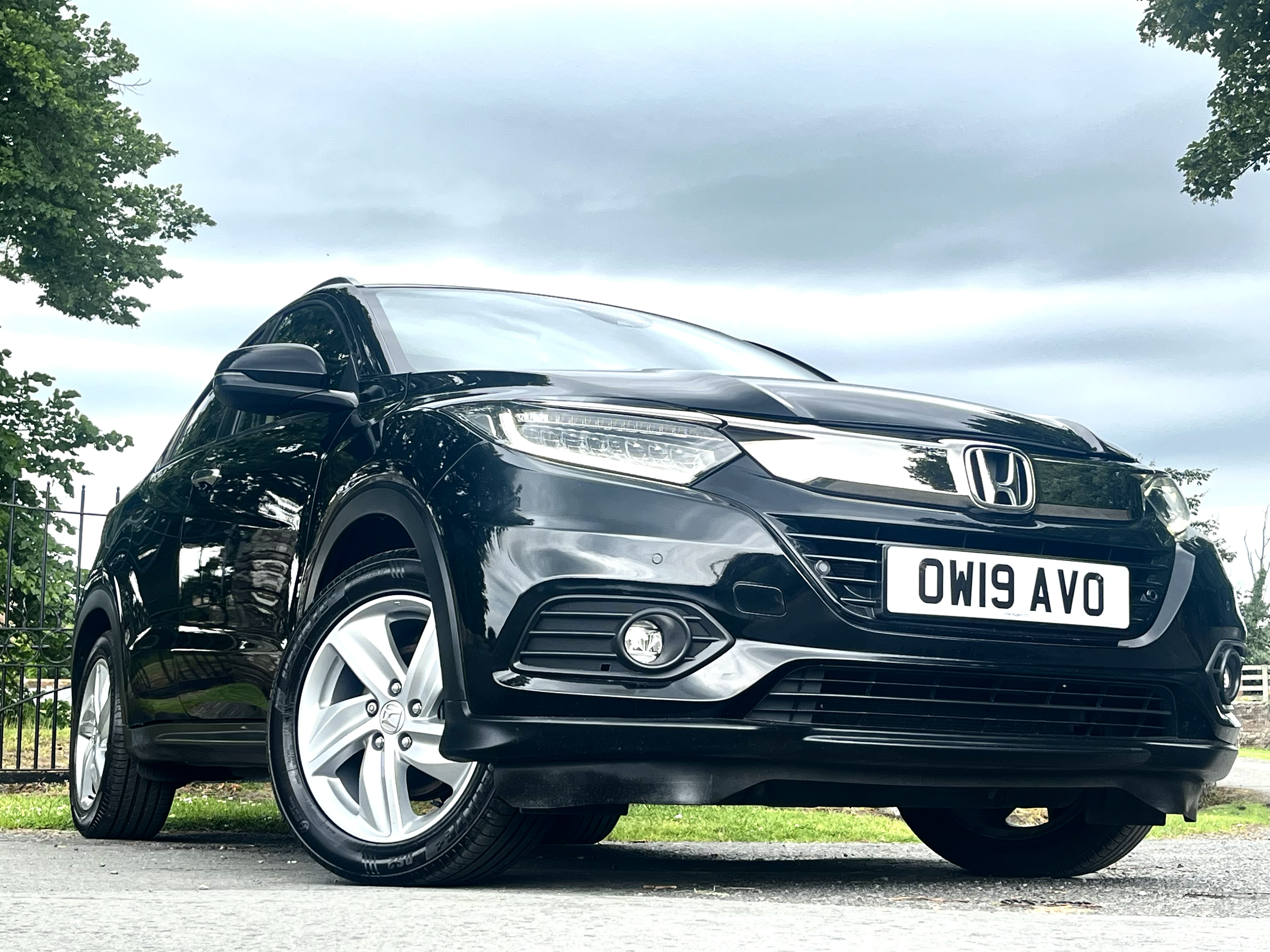 A black Honda car parked outdoors on a street, with green trees and a cloudy sky in the background.