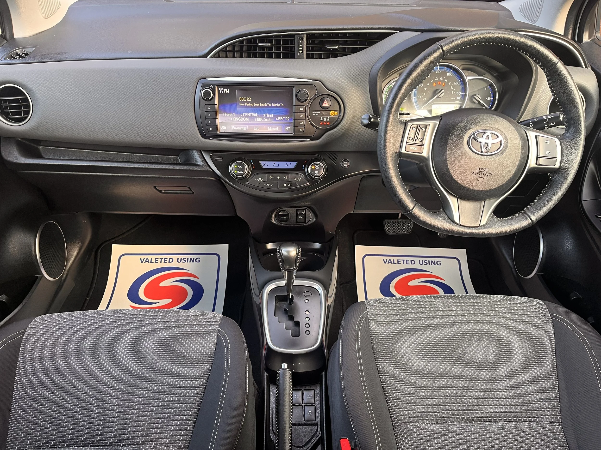 Interior of a Toyota car showing the dashboard, steering wheel, gear shift, and front seats with floor mats.