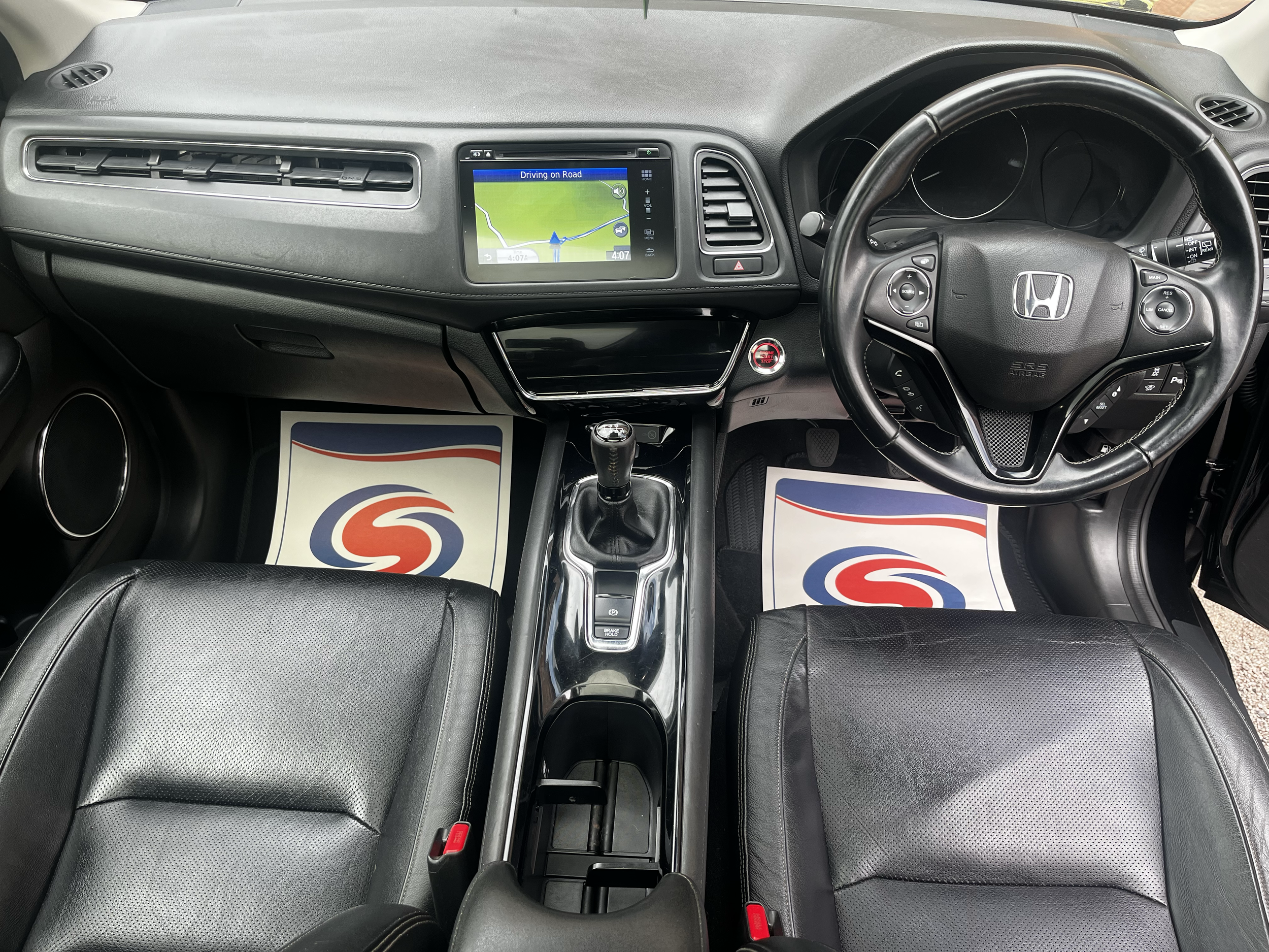 View of the interior of a Honda vehicle dashboard, including steering wheel, GPS screen showing a map, and gear shift, with black leather seats and floor mats.