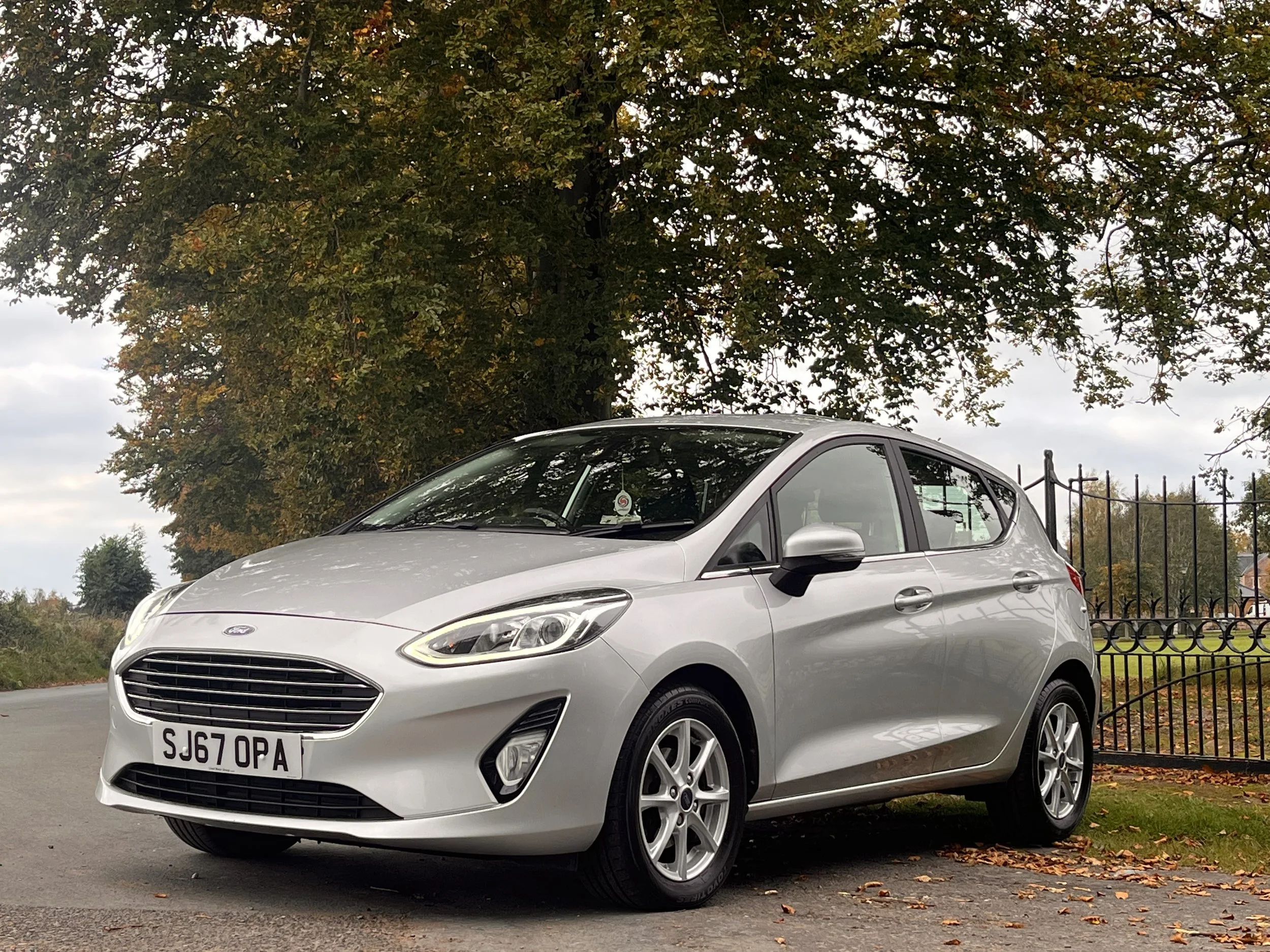 Silver Ford Fiesta parked on the side of a road with autumn trees and fence in the background.
